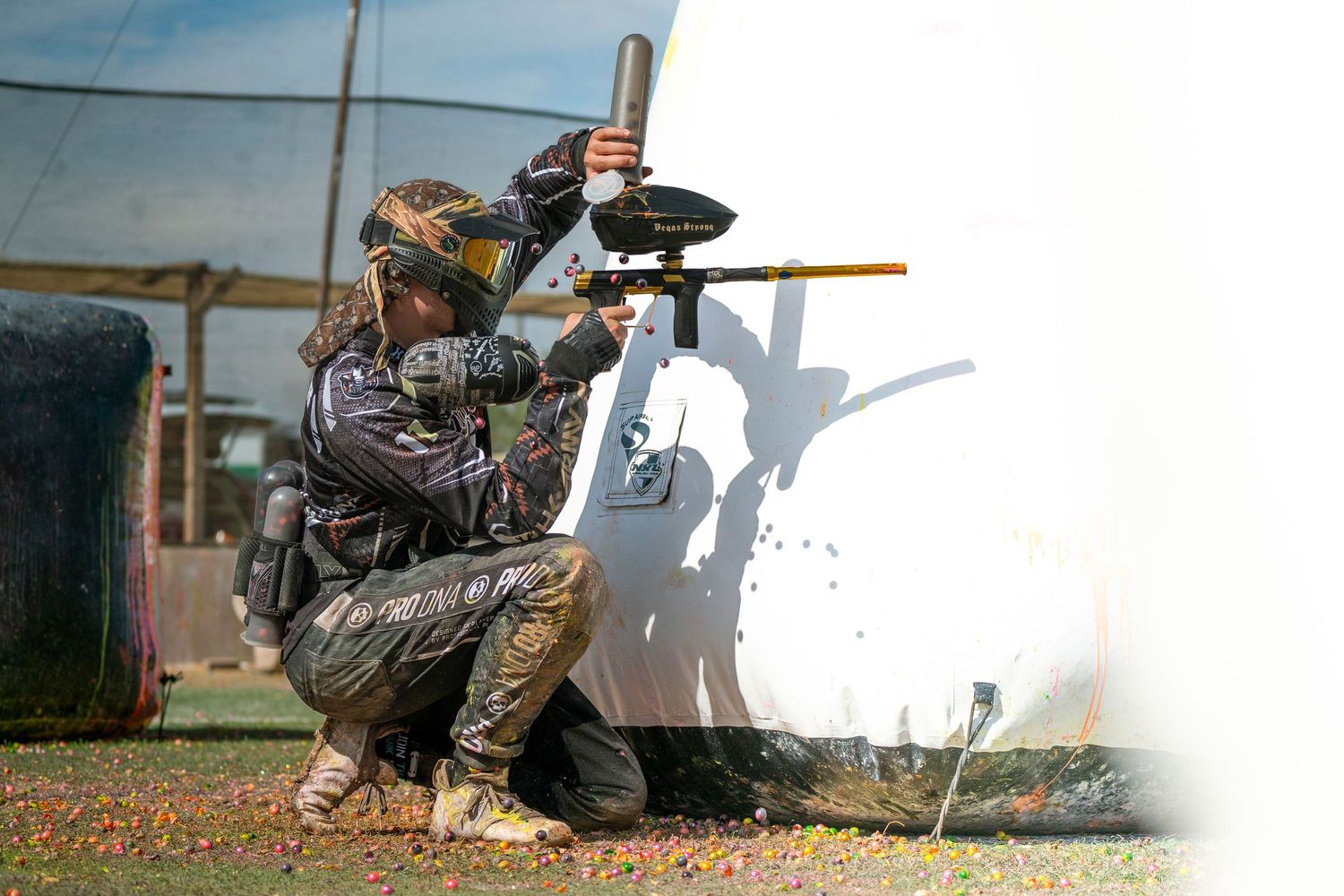 Tactical paintball player in camo gear crouching behind white bunker during match.
