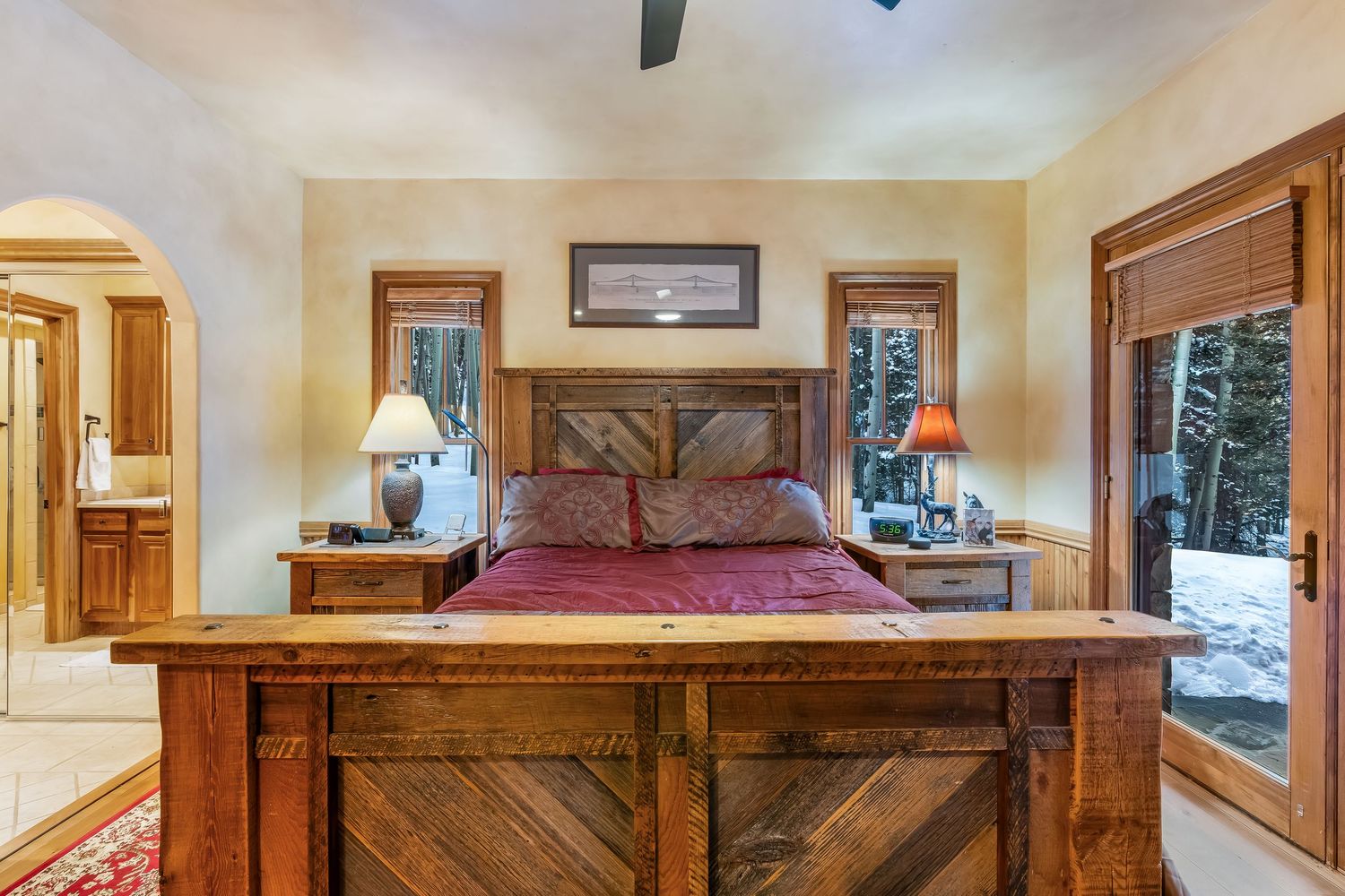 Luxurious mountain lodge bedroom with rustic wood furnishings, stone fireplace, and panoramic windows overlooking snowy scenery.