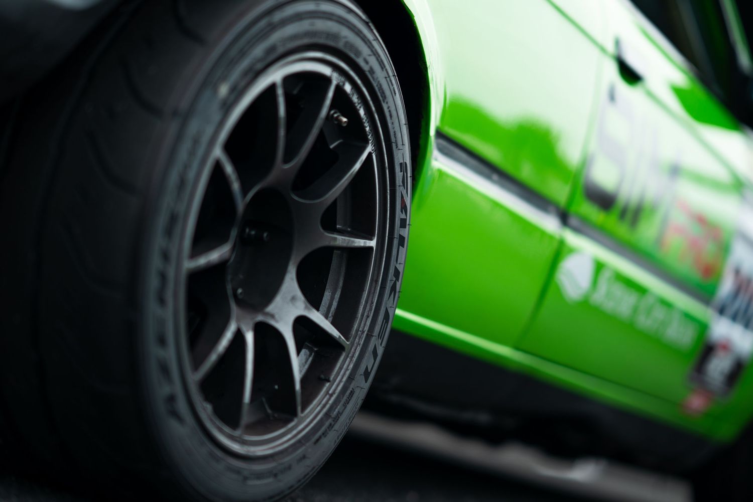 Close-up of black alloy wheel and tire on a green race car at a motorsport event.