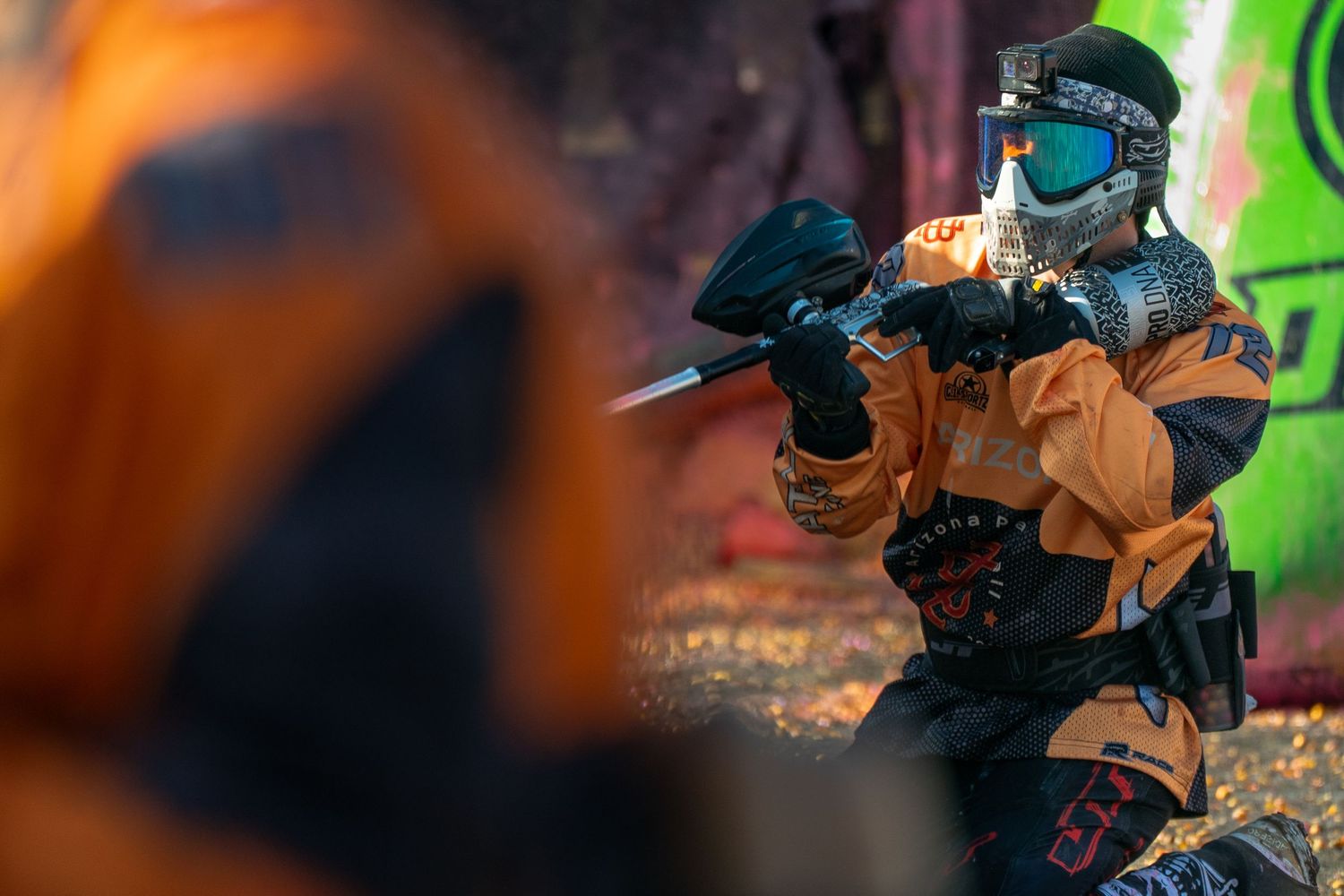 Player in orange paintball gear aims marker during competitive paintball match outdoors.
