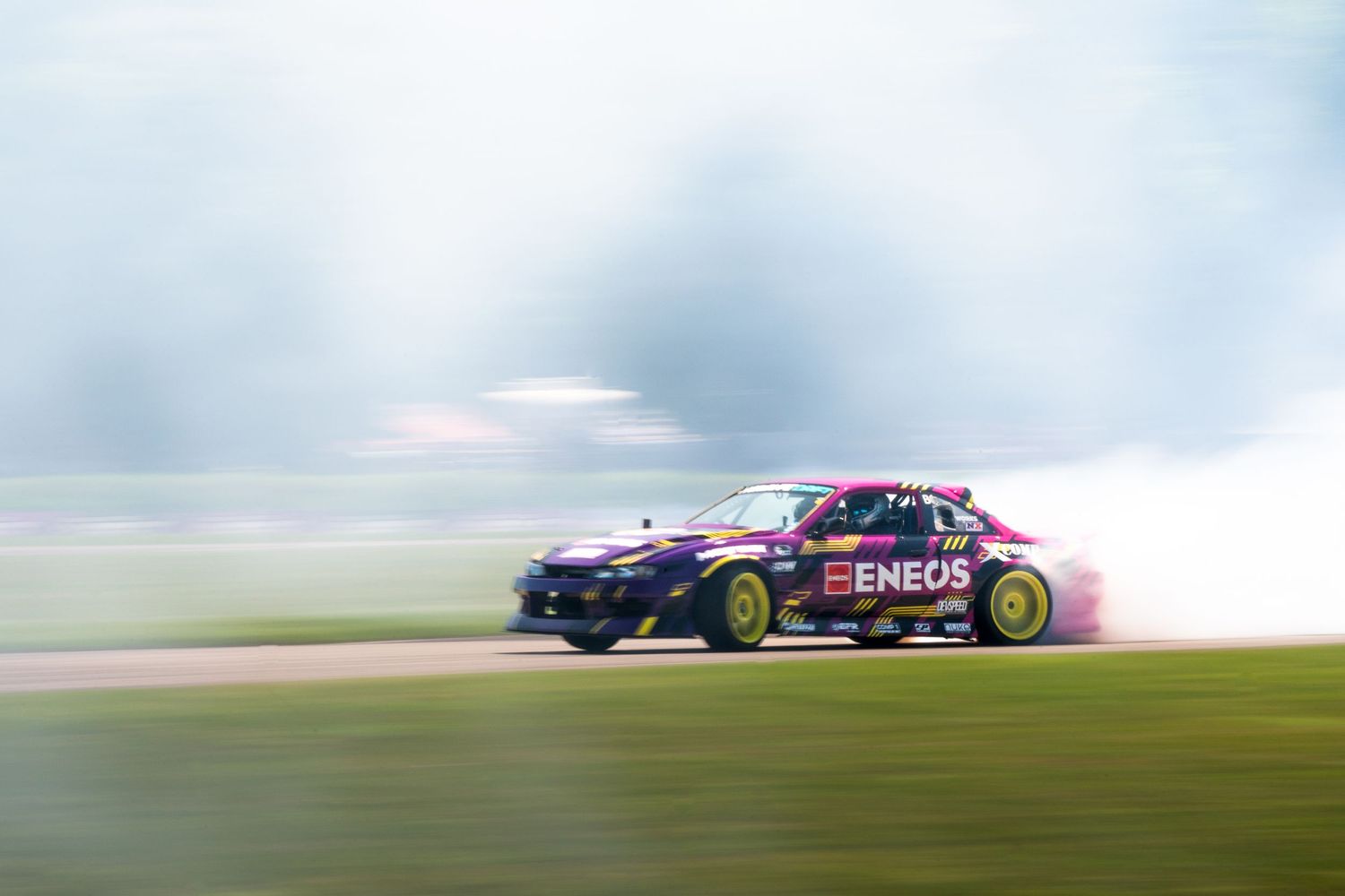 A racing car with ENEOS sponsorship drifting on a track, creating thick smoke clouds during a motorsport event.