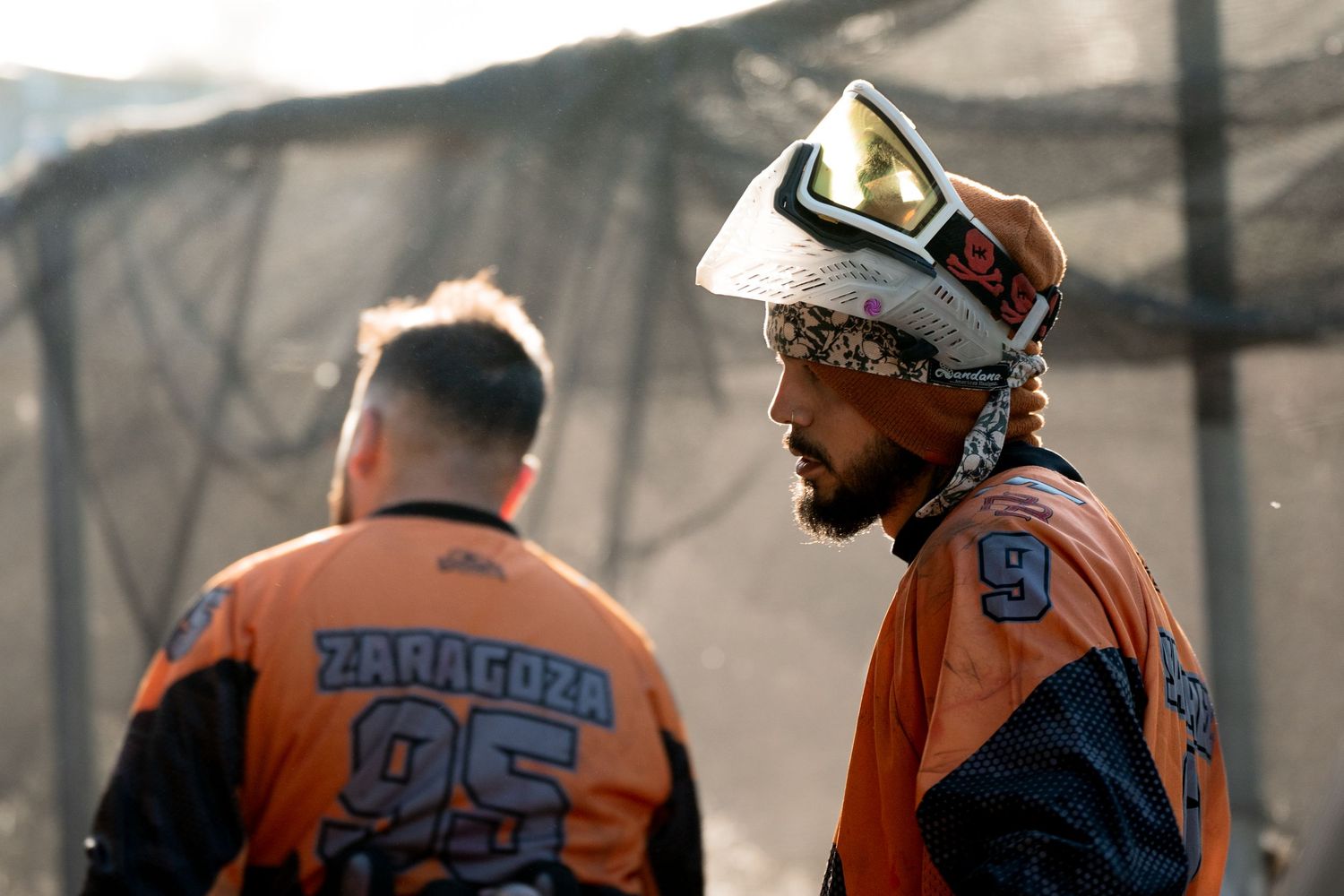 Two paintball players in orange Zaragoza jerseys wearing protective masks stand on a paintball field.