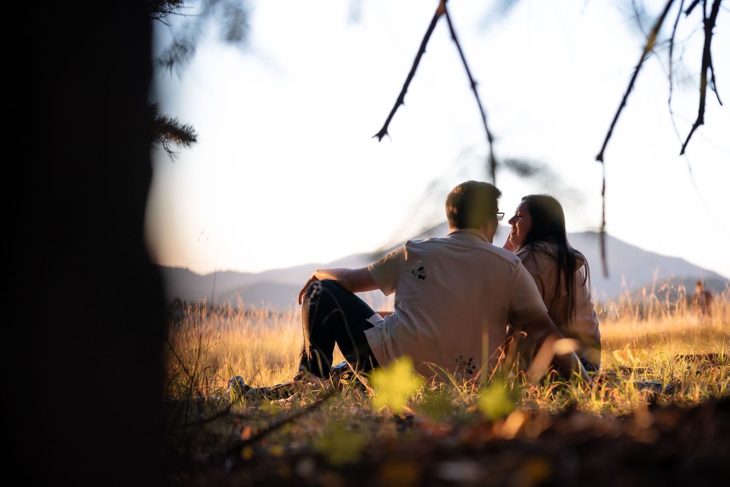 Couple sitting close together on a grassy hillside at sunset, gazing toward distant mountains beneath tree branches.