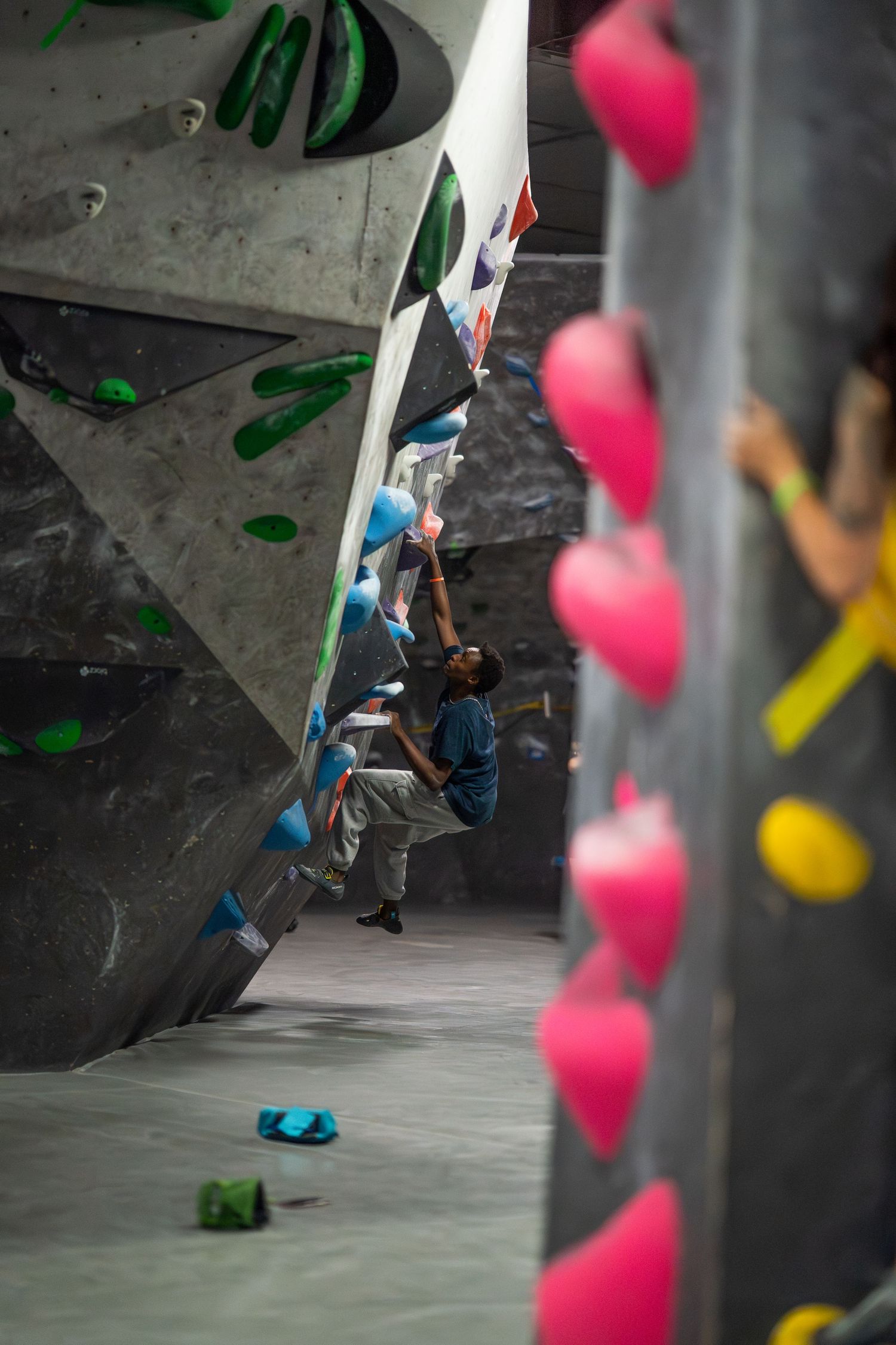Climber in a blue shirt reaches for holds on an angled indoor bouldering wall with green and pink grips.