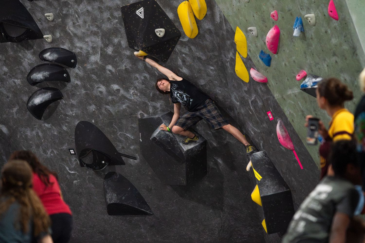 Climber in a dark shirt stretches across a black volume on a textured indoor bouldering wall with colorful holds as spectators watch nearby.