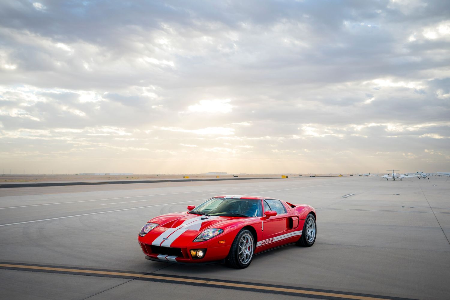 Red Ford GT supercar parked on an airport runway against a dramatic cloudy sky at sunset.