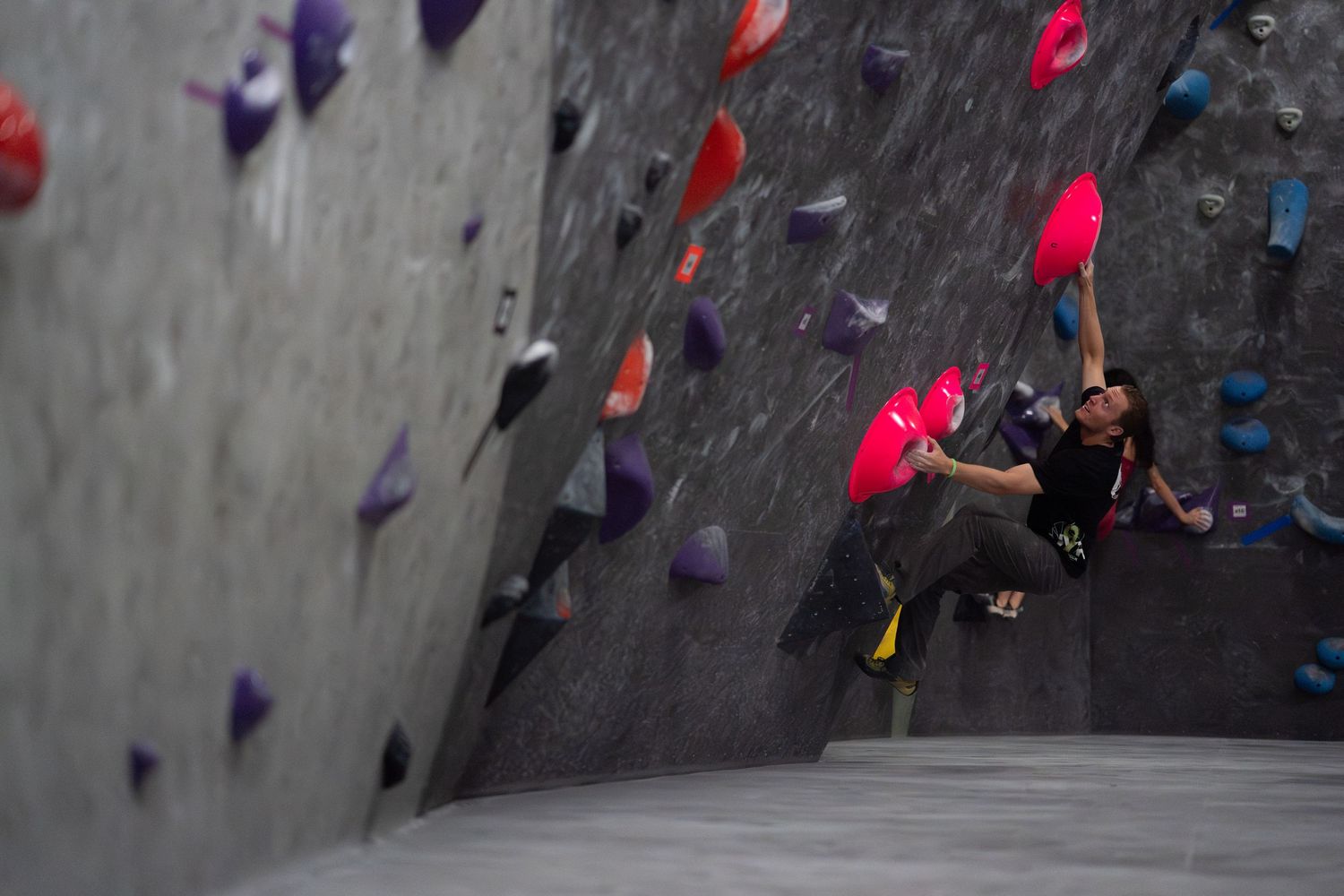 Climber in dark clothing reaches for a bright pink hold on an overhanging indoor bouldering wall covered in colorful climbing holds.