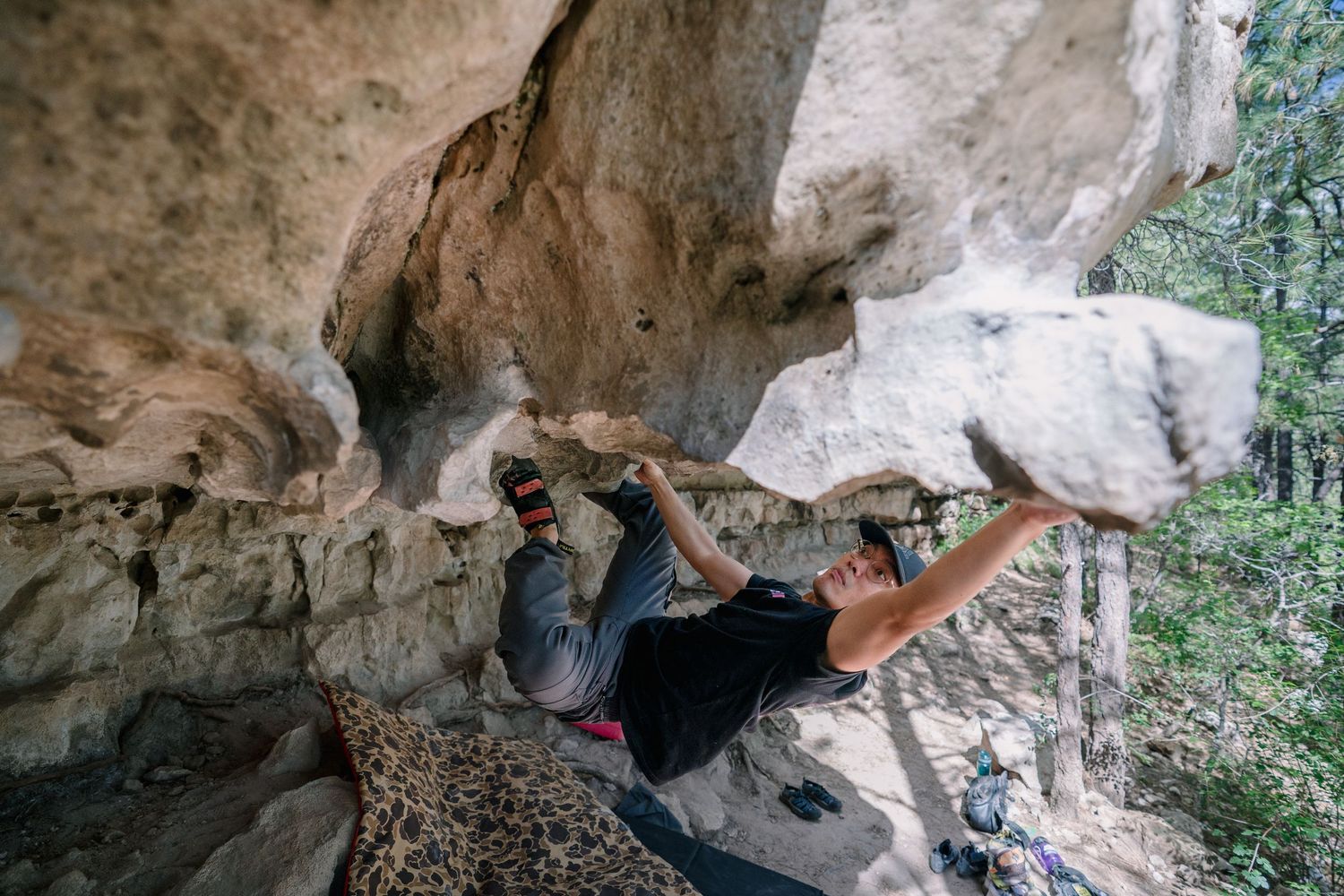 A climber in a black shirt stretches across a steep sandstone overhang while bouldering outdoors in a forested area.