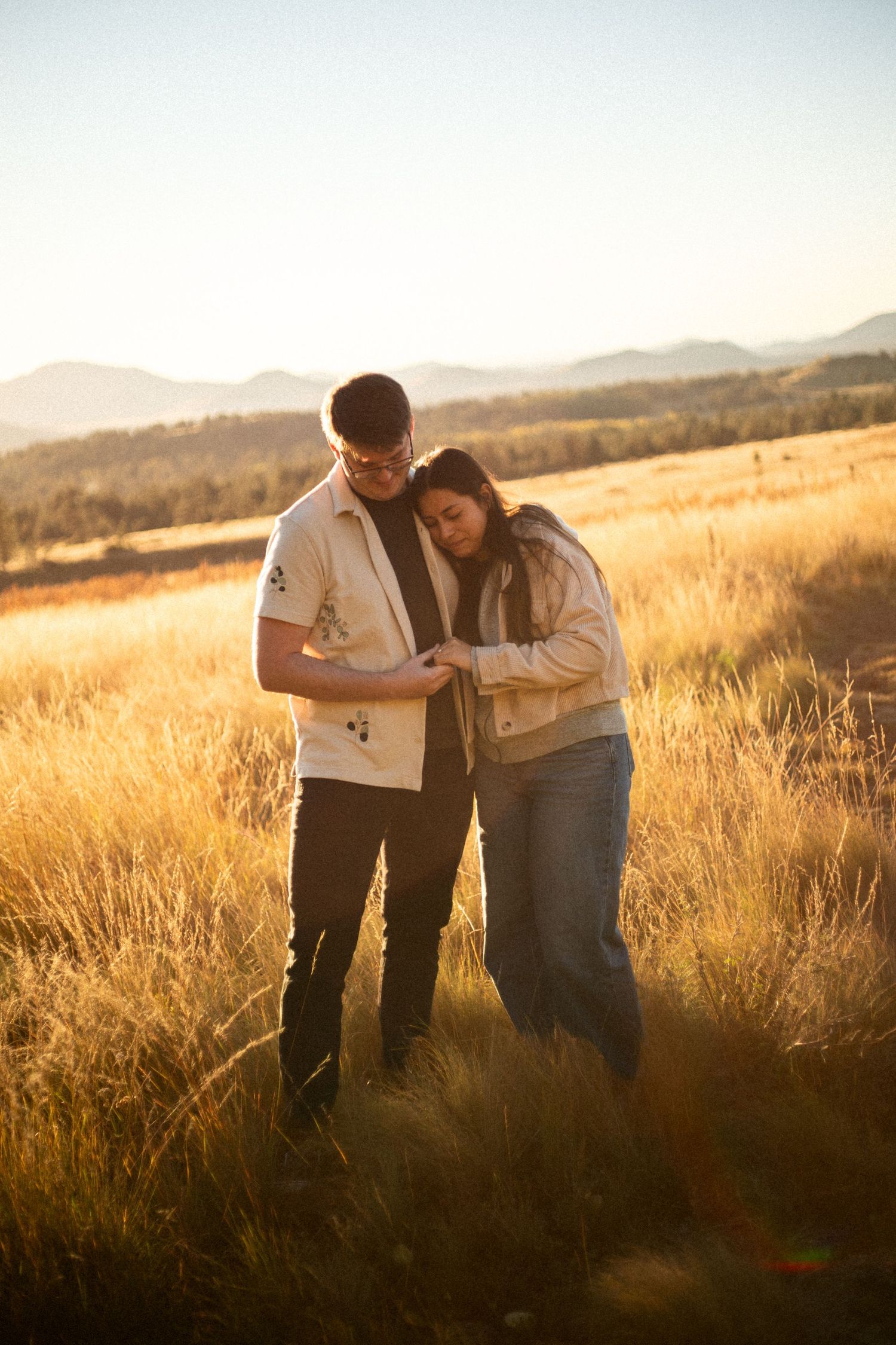 Couple embracing in a golden grassy field at sunset, with rolling hills and a soft hazy sky in the background.