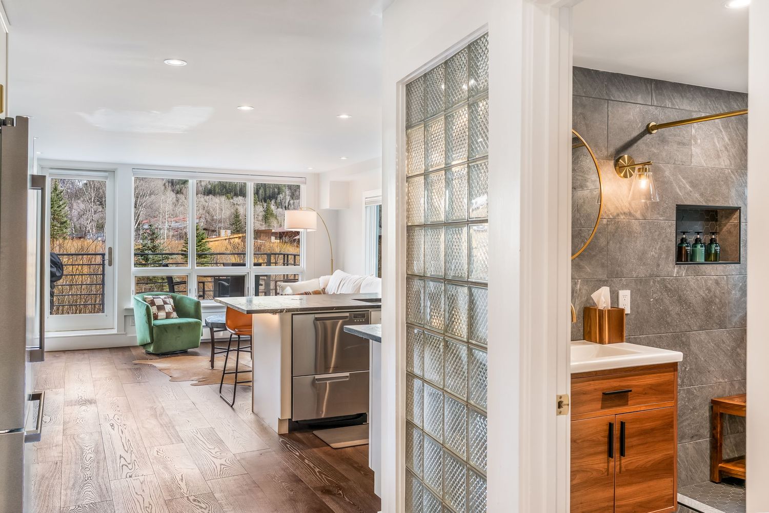 Modern open-concept kitchen with glass block wall, hardwood floors, and bright sunlit living area beyond.