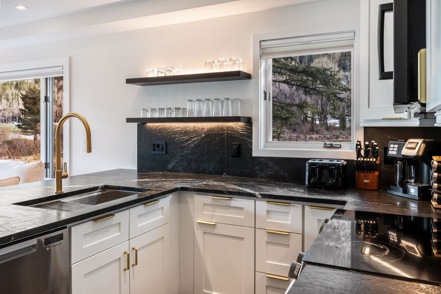 Modern kitchen with black granite countertops, white cabinets, gold hardware, floating shelves, and a gold faucet near a bright window.