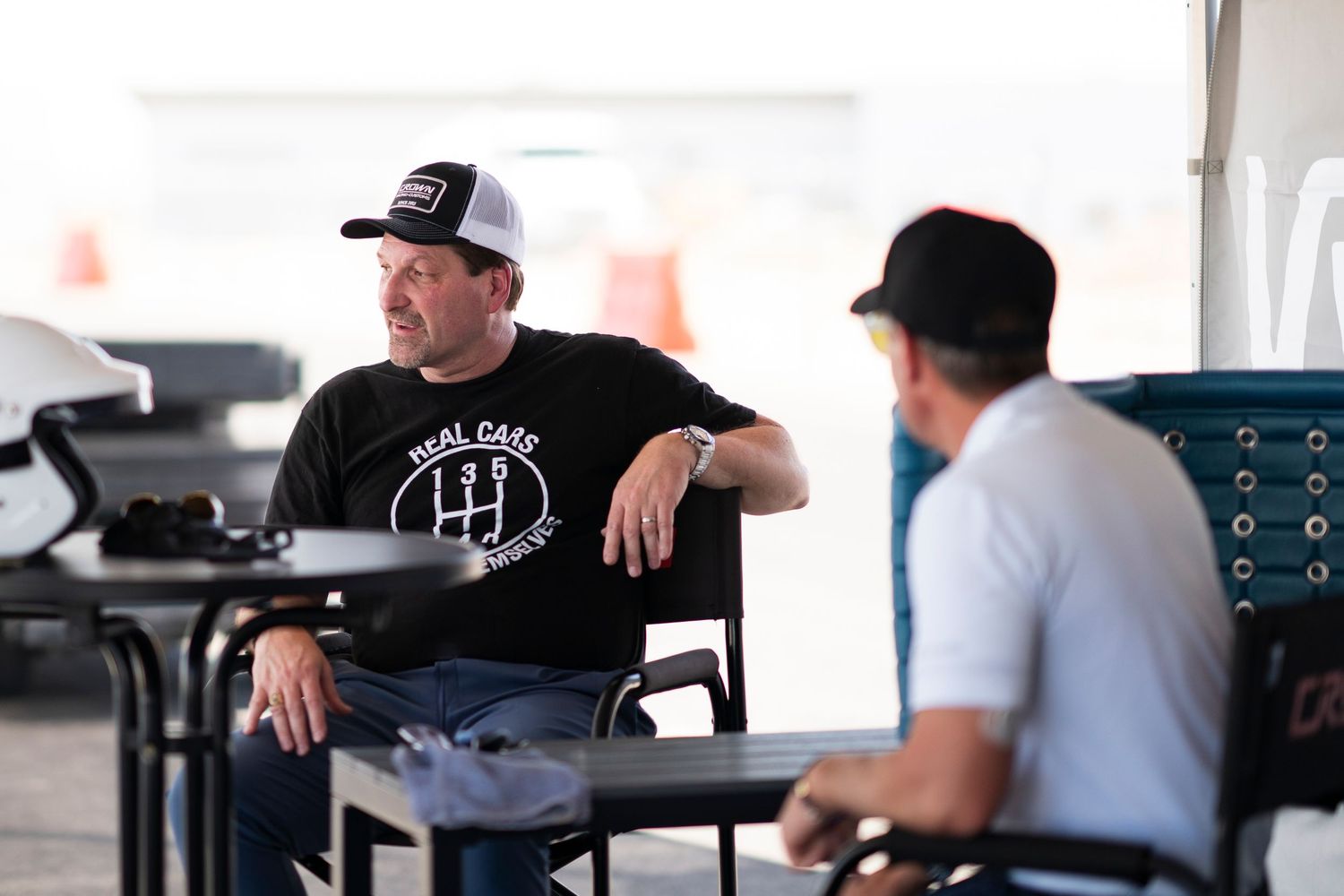 Two people in baseball caps having a conversation while seated in a covered outdoor area.
