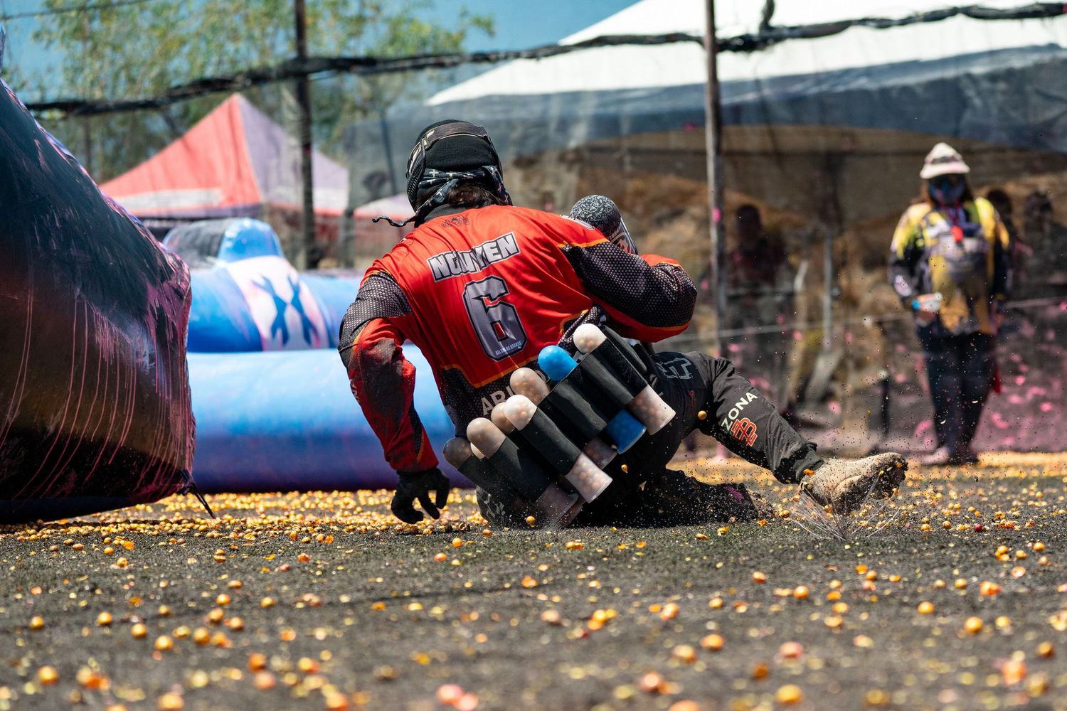 Paintball player in red jersey crouches low behind bunker during intense outdoor tournament competition.