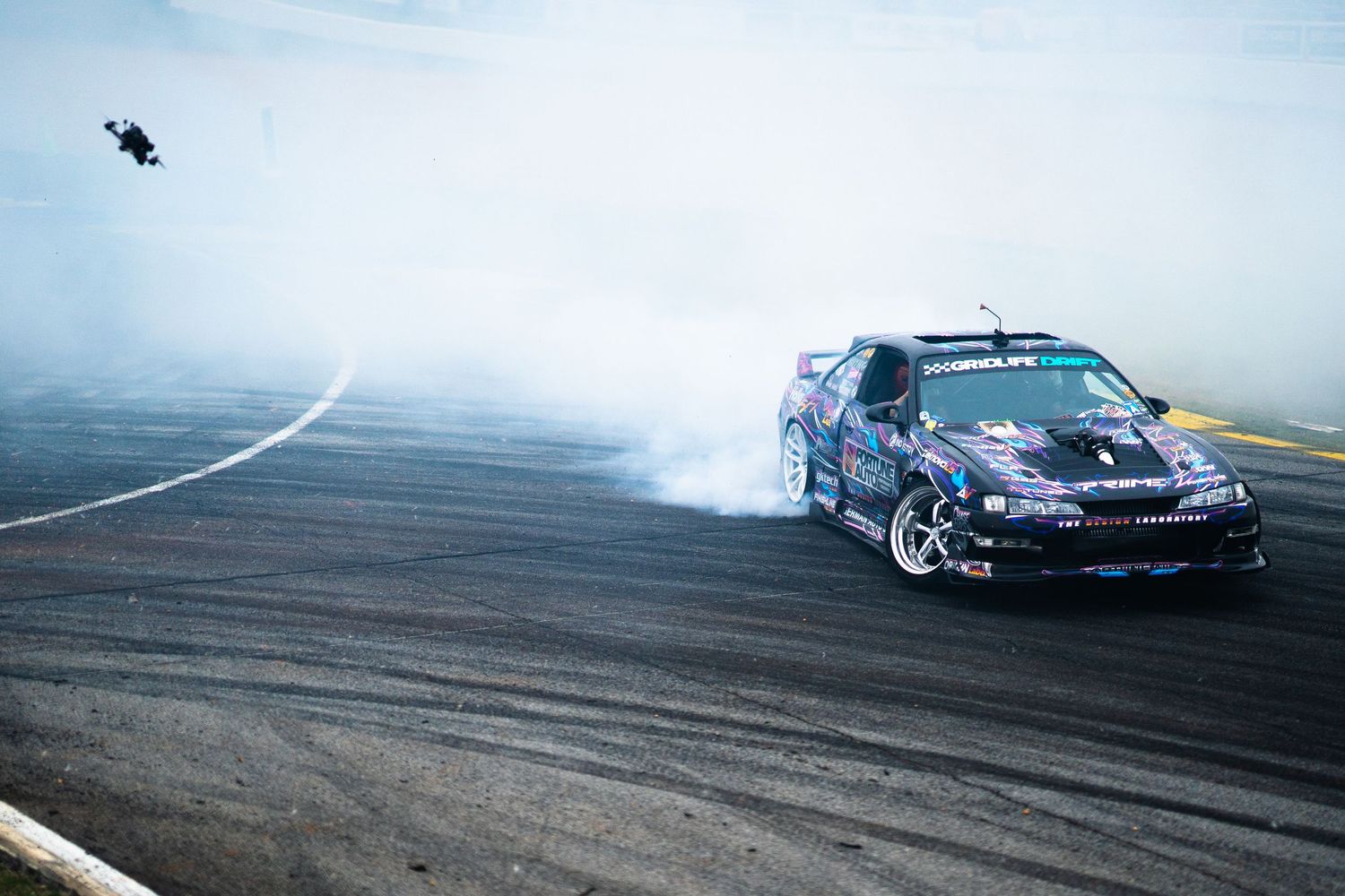 Dark purple drift car creates massive tire smoke clouds while drifting on a motorsport track.