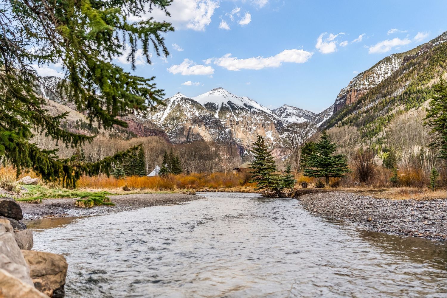 Scenic mountain river landscape with snow-capped peaks, evergreen trees, and golden autumn foliage under blue sky.