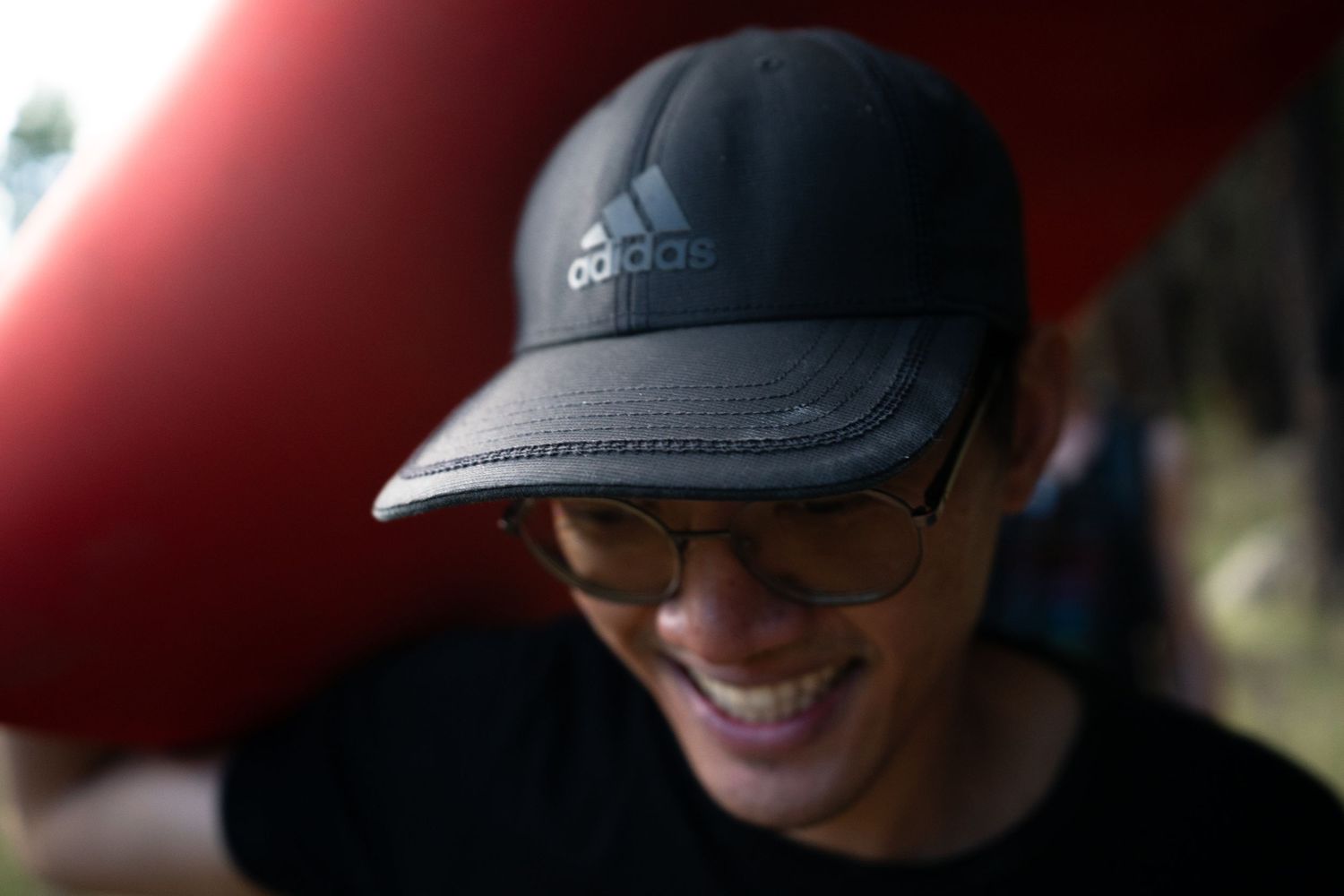 Smiling person wearing round glasses, a black Adidas cap and a black t-shirt, looking down with a blurred red object and greenery behind.
