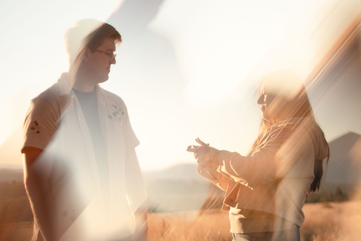Two people stand facing each other in a golden grassy field at sunset, captured in a dreamy double exposure with warm light flares.
