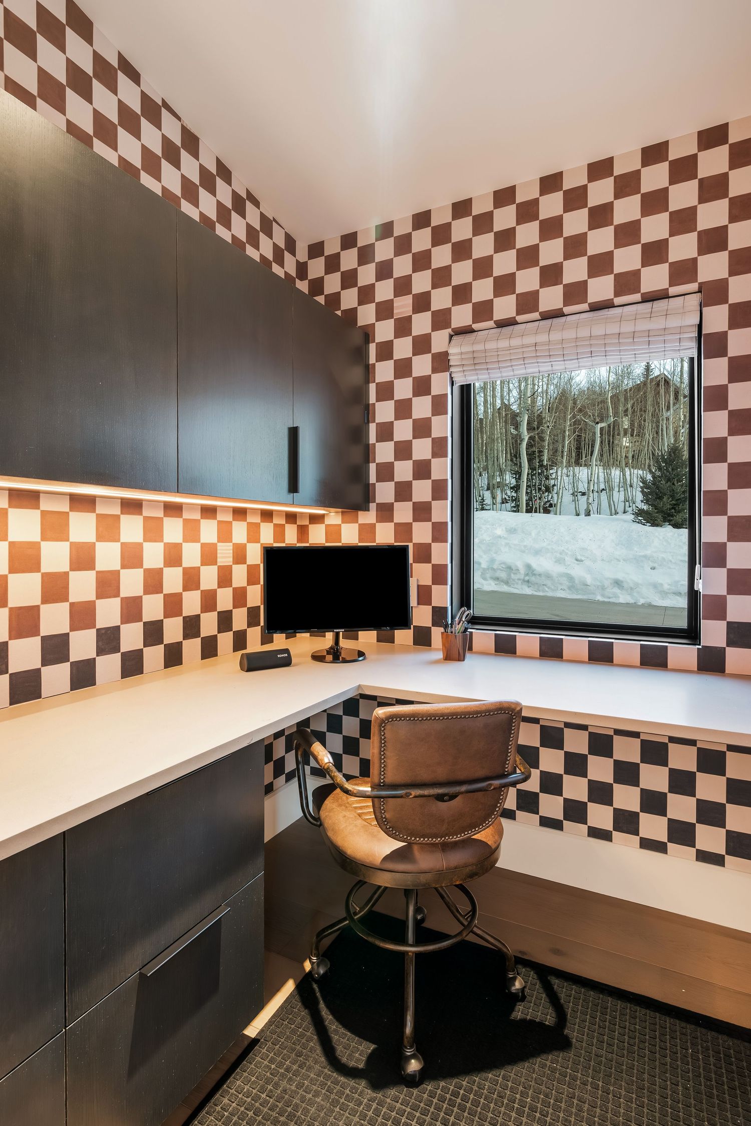 Home office with brown and white checkered walls, black cabinets, and large window showing winter scenery.