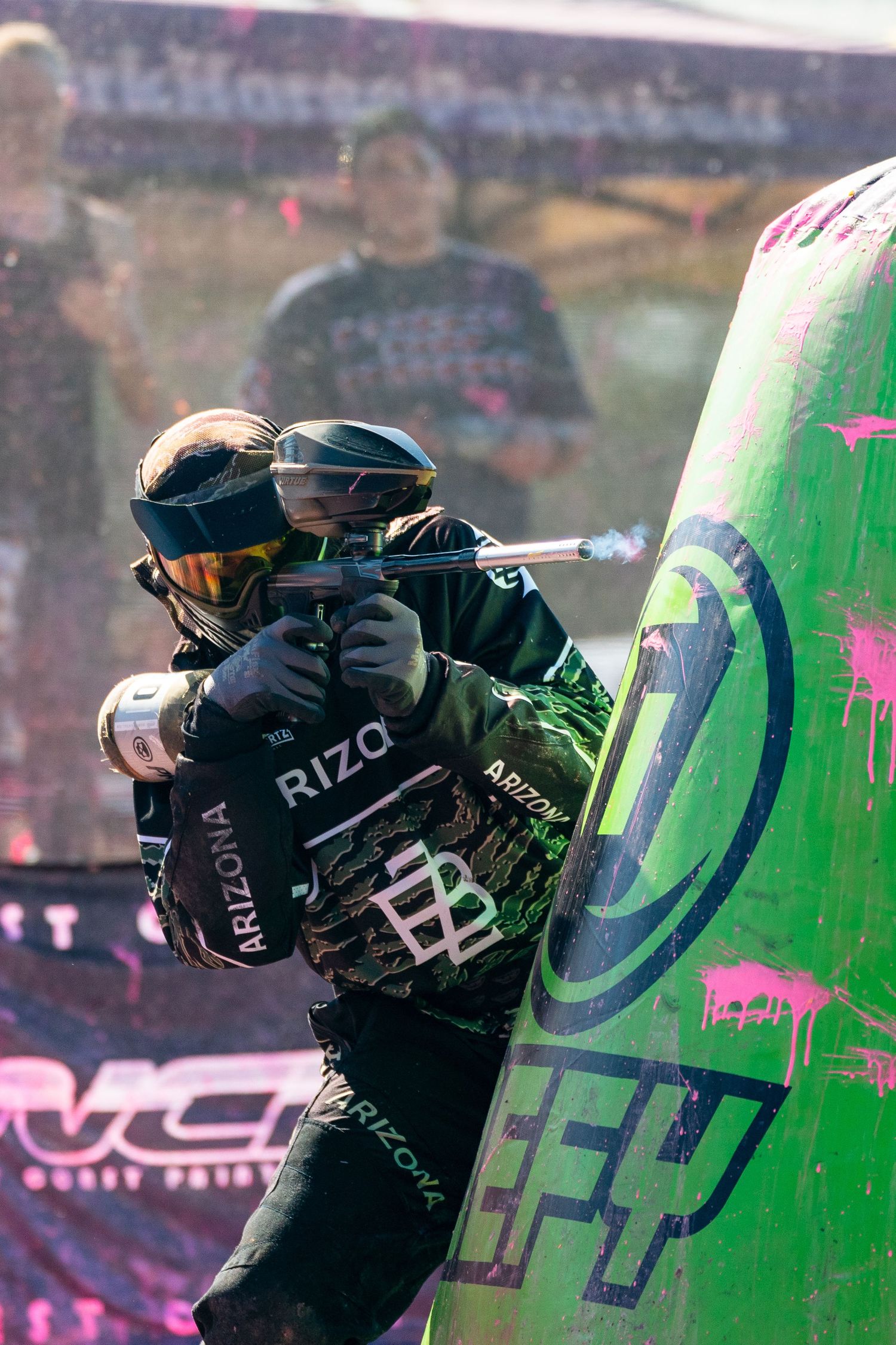 Arizona paintball player aims marker behind green inflatable bunker during competitive speedball tournament match.