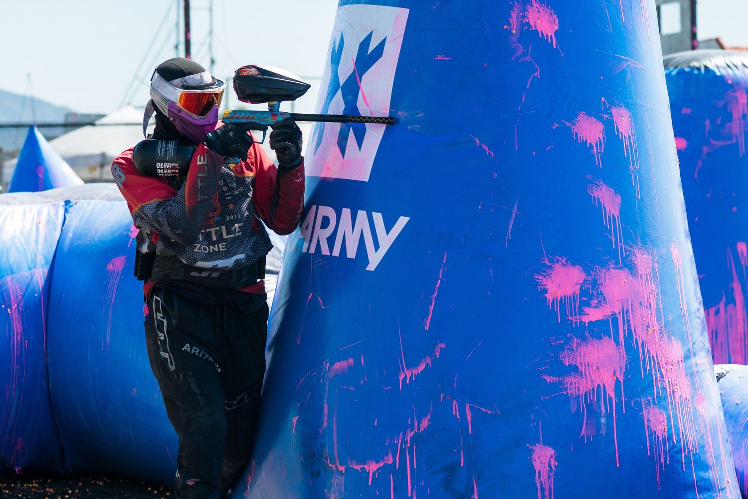 A paintball player in red gear aims their marker behind a blue inflatable bunker with pink paint splatters at an outdoor tournament.