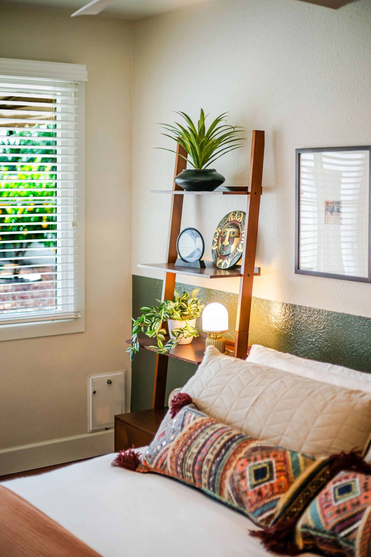 Cozy bedroom interior with decorative pillows, houseplants, and natural light streaming through white plantation shutters.