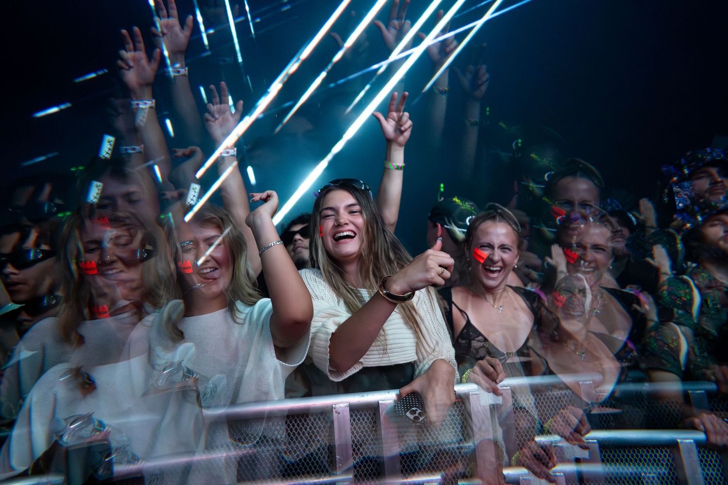Excited crowd of fans cheering and raising hands at a vibrant nighttime music festival with laser lights.