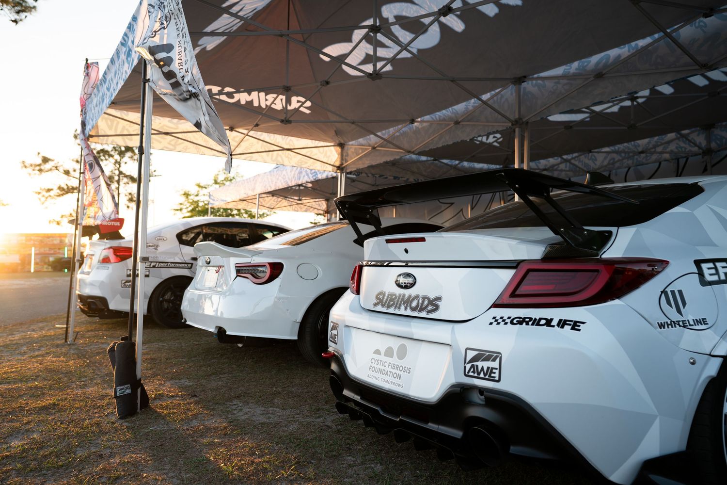 White modified sports cars lined up under event tents at a car show, featuring large rear spoilers and custom body kits.