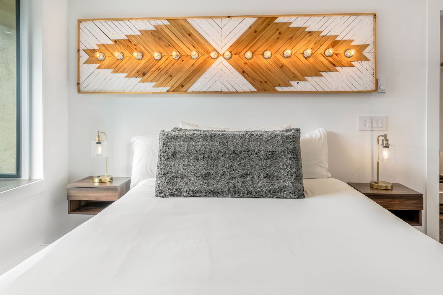 Modern bedroom with white bedding, wood nightstands, and a decorative geometric wood wall art above the bed.