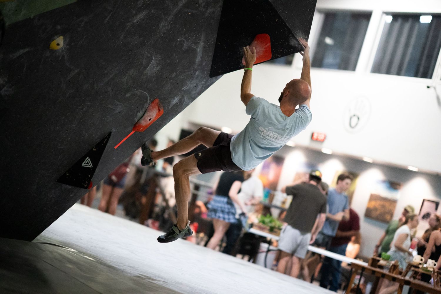 Climber in a gray t-shirt reaches for a hold on an overhanging bouldering wall inside an indoor gym while spectators watch below.