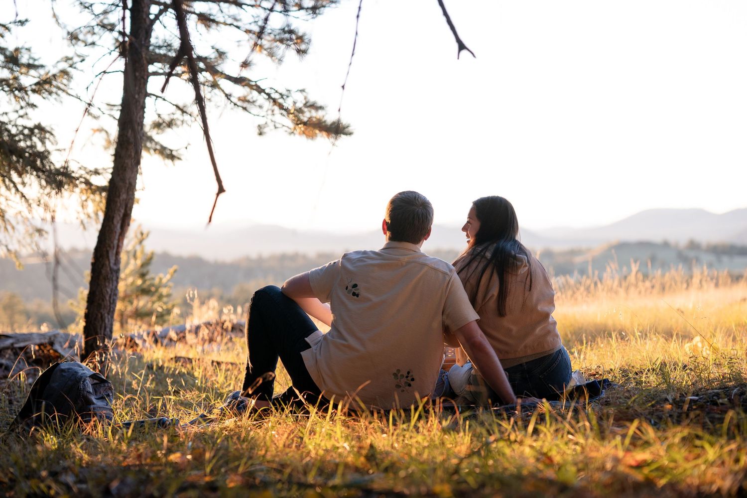 Couple sitting together on the grass beneath a pine tree, gazing at a mountain view in warm evening light.