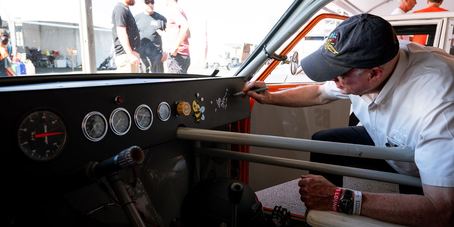 Multiple vintage race cars are lined up in a row with drivers seated in their cockpits during a racing event.