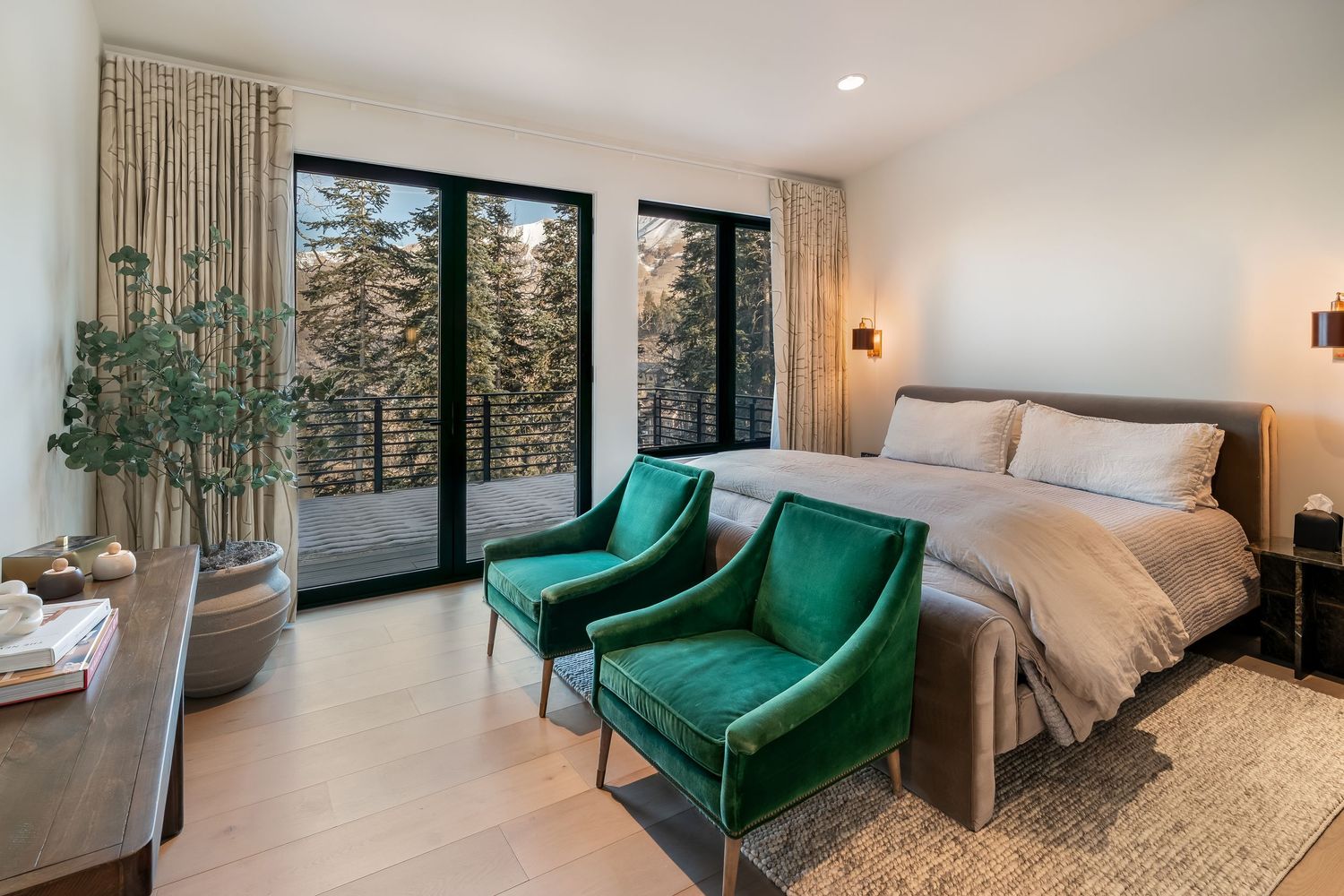 Modern bedroom with floor-to-ceiling windows, emerald green velvet chairs, and mountain views beyond the balcony.