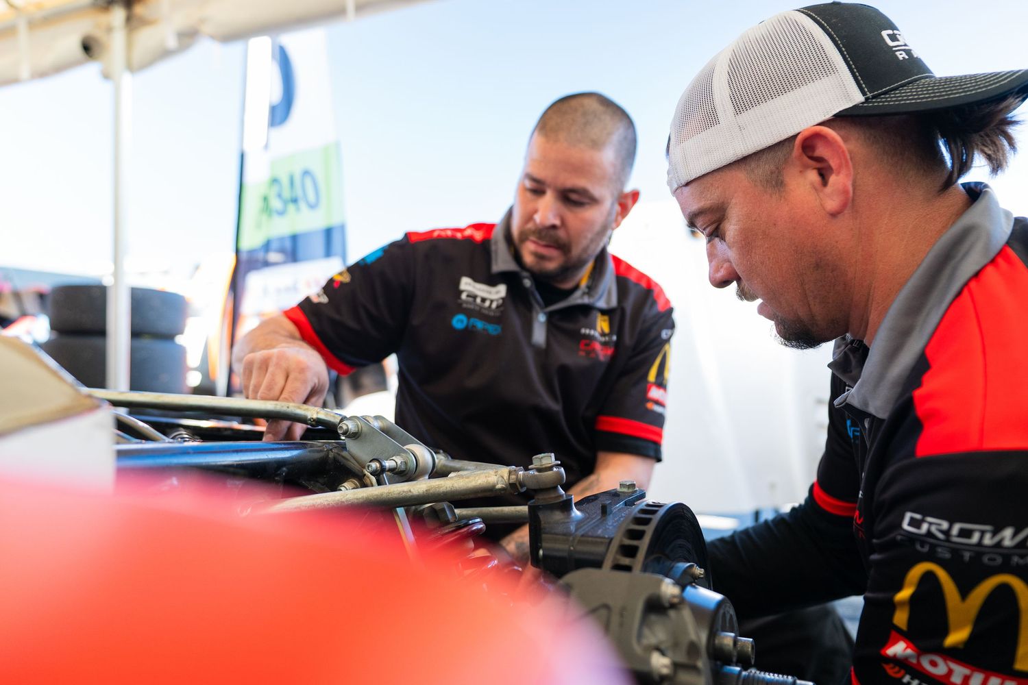 Pit crew members in racing uniforms collaborate on vehicle preparations in a covered service area.