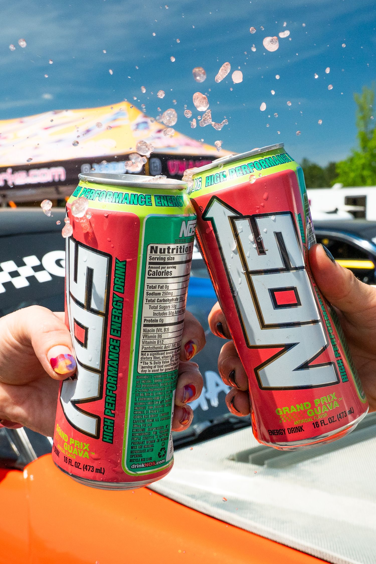 Two hands clinking NOS High Performance Energy Drink cans at a racing event with liquid splashing.