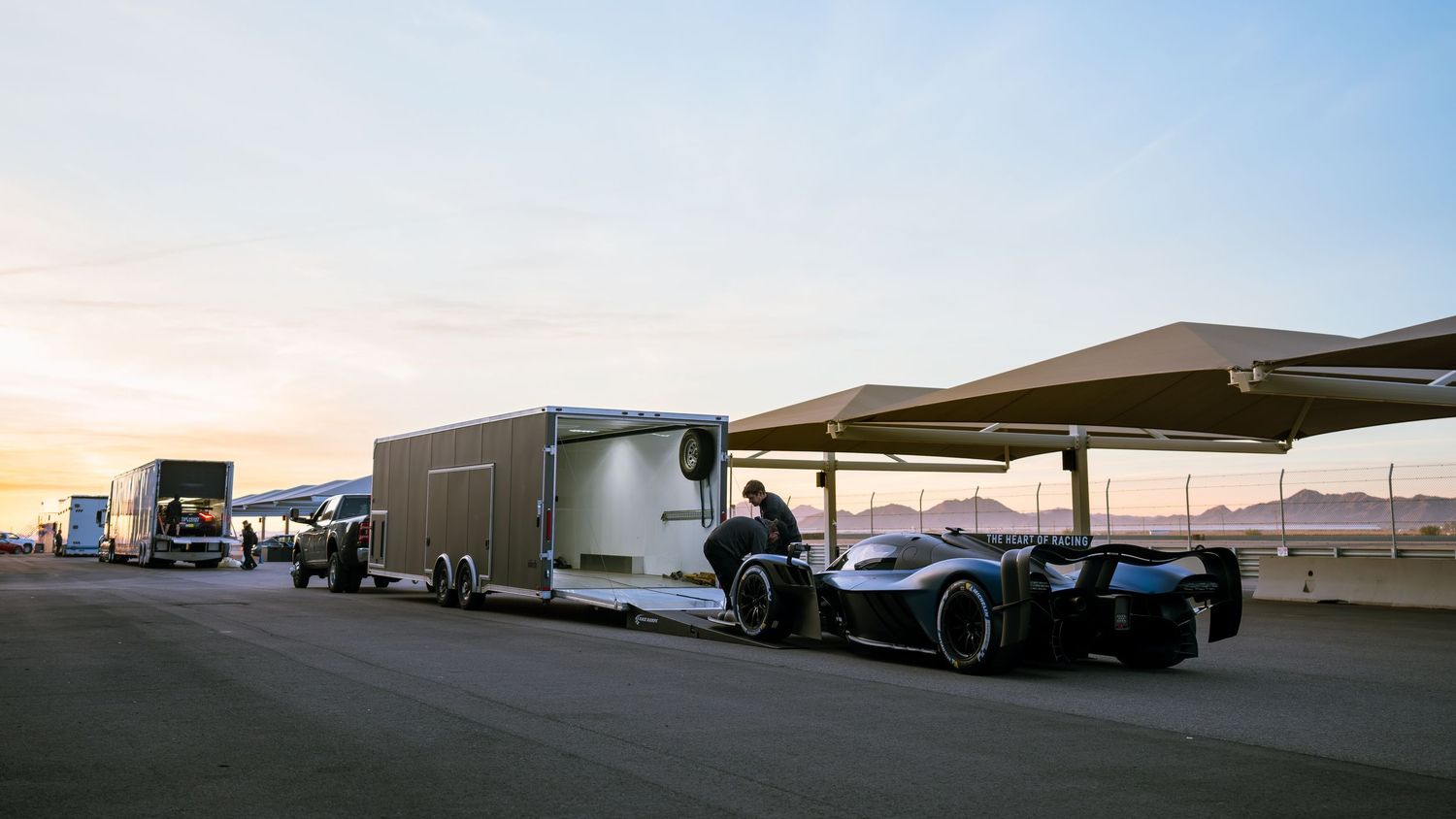 Race car transporter and haulers parked at sunset with a sleek performance vehicle under a canopy.