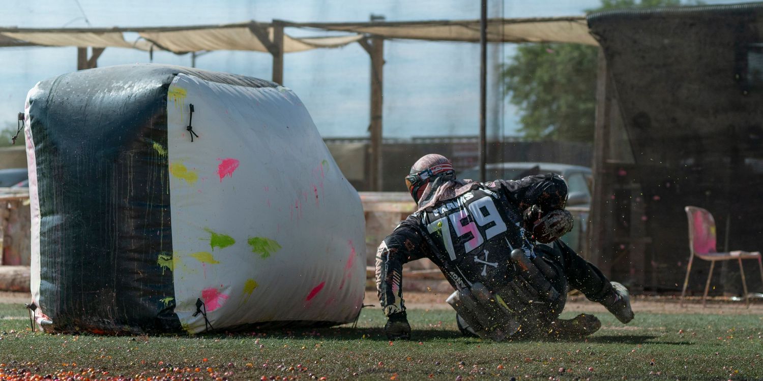 Player in tactical gear takes cover behind inflatable bunker during paintball match.