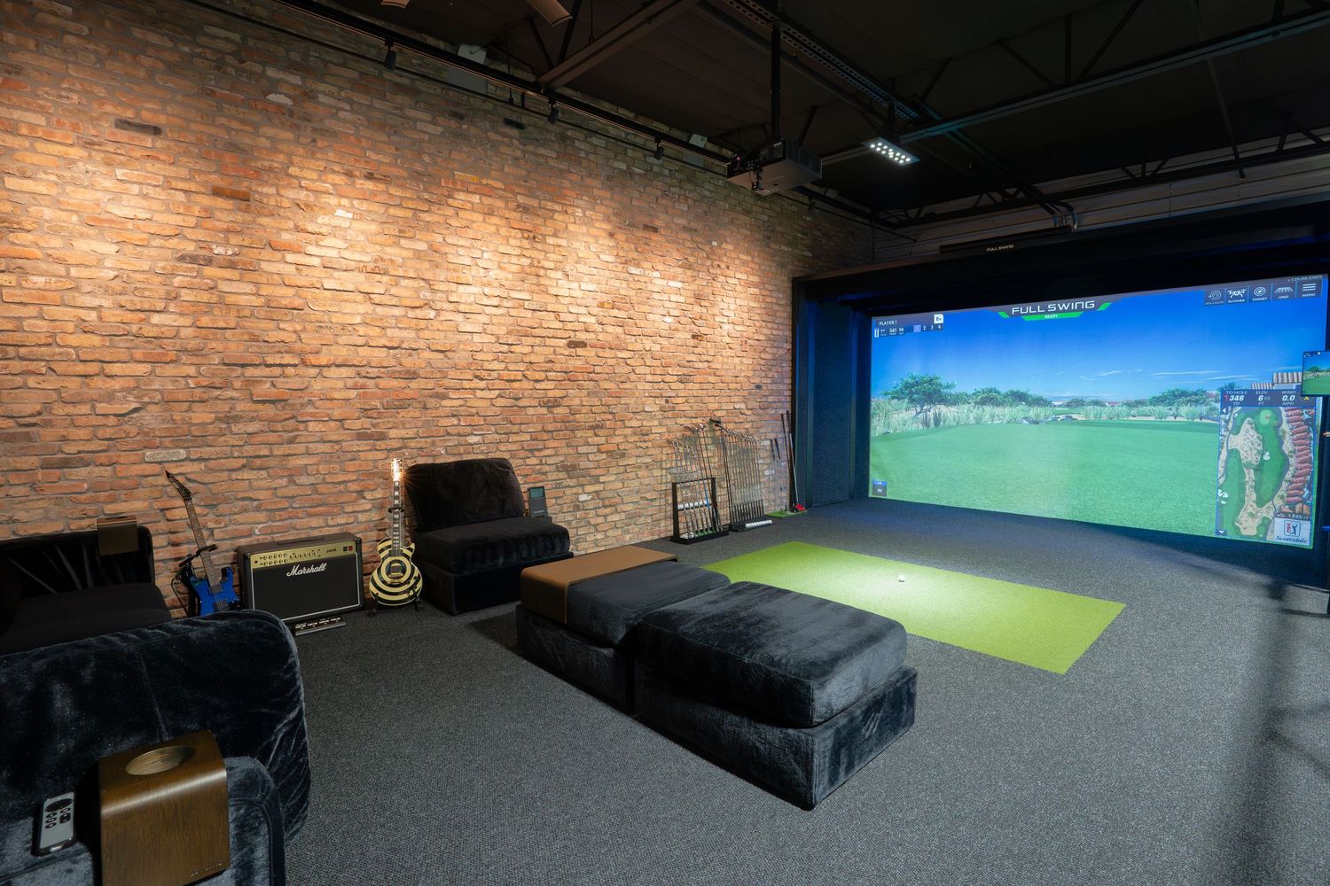 Indoor golf simulator setup with projection screen and comfortable lounge seating against brick wall.