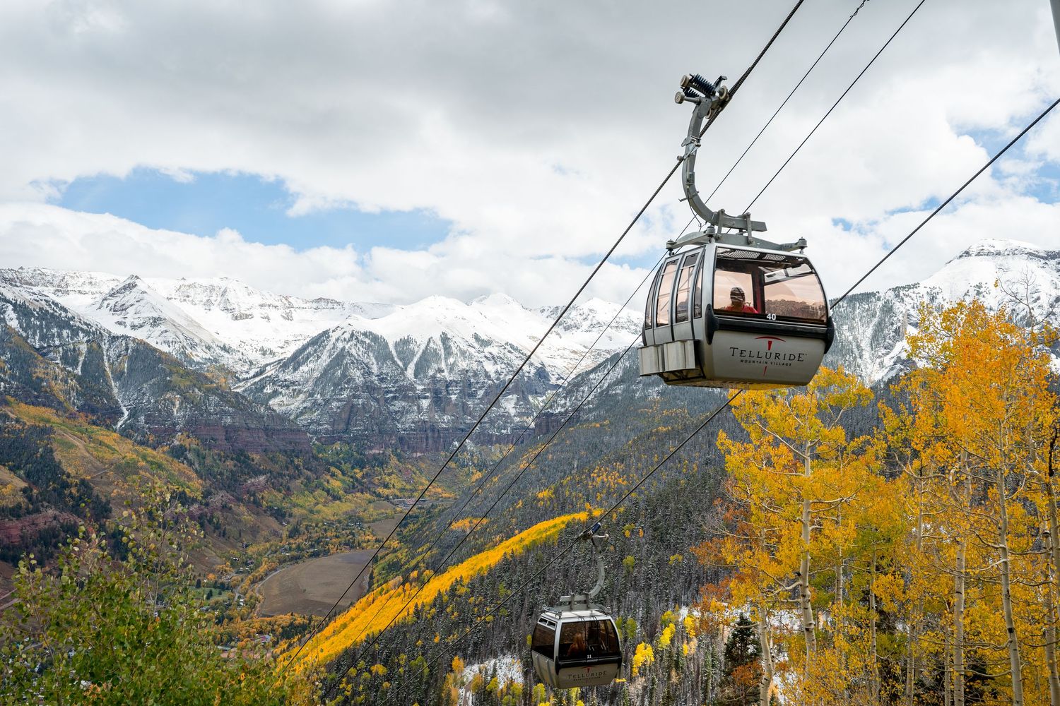Gondola lift cars ascending snow-capped mountains with golden fall foliage in the foreground.