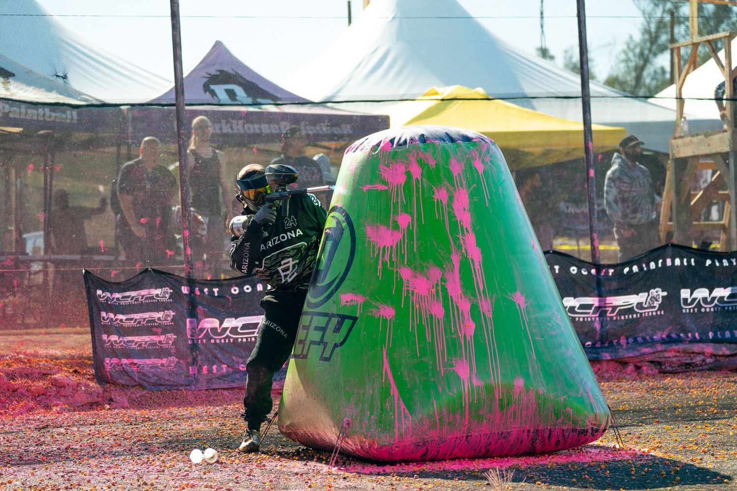 Paintball player crouching behind a colorful inflatable bunker during an outdoor paintball tournament match.