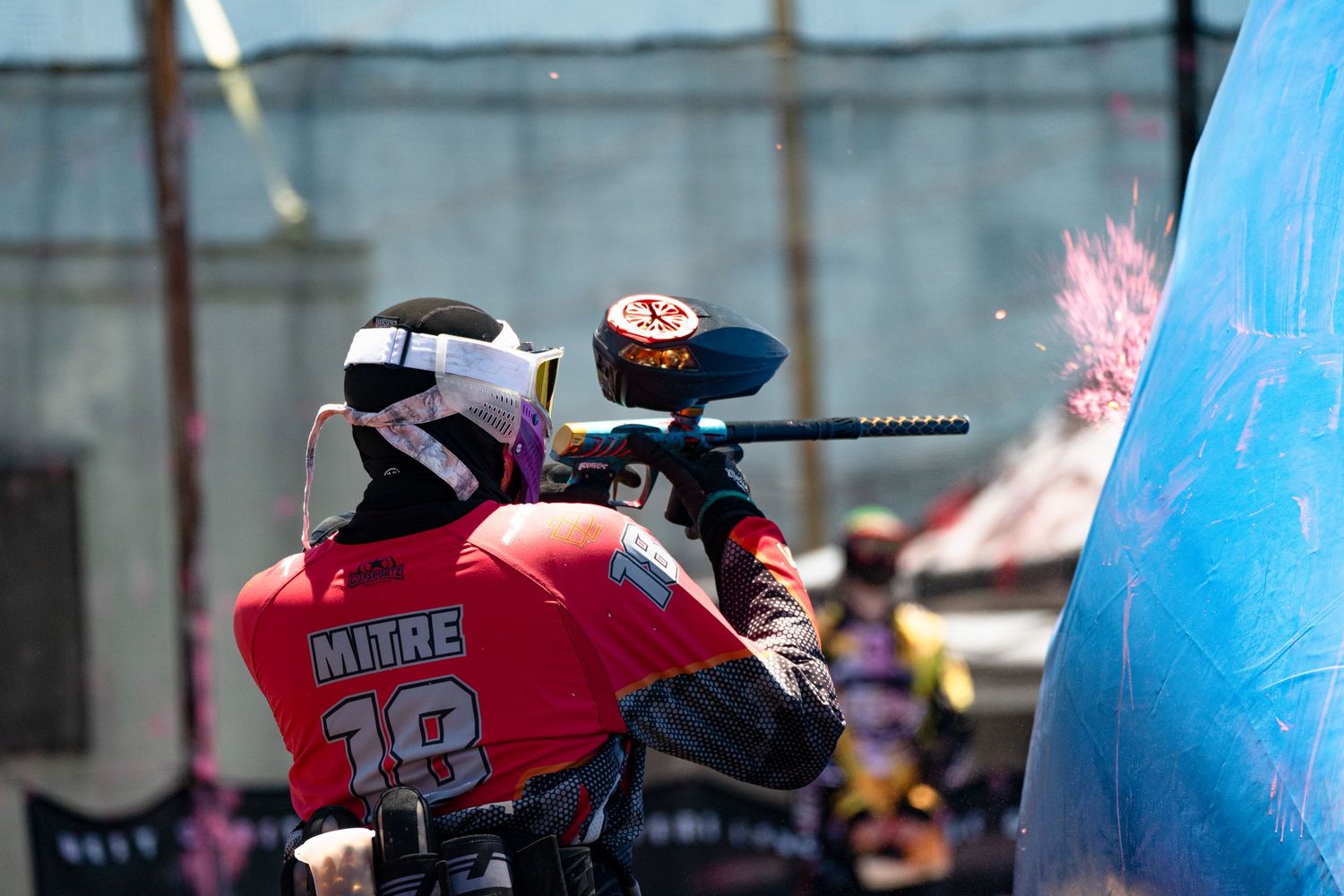 Paintball player wearing Mitre jersey number 18 fires marker during intense outdoor tournament competition.