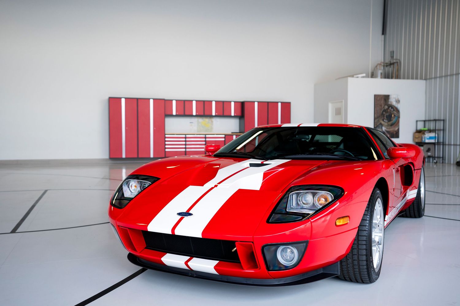 A red Ford GT sports car with white racing stripes parked in a pristine garage with red storage cabinets.