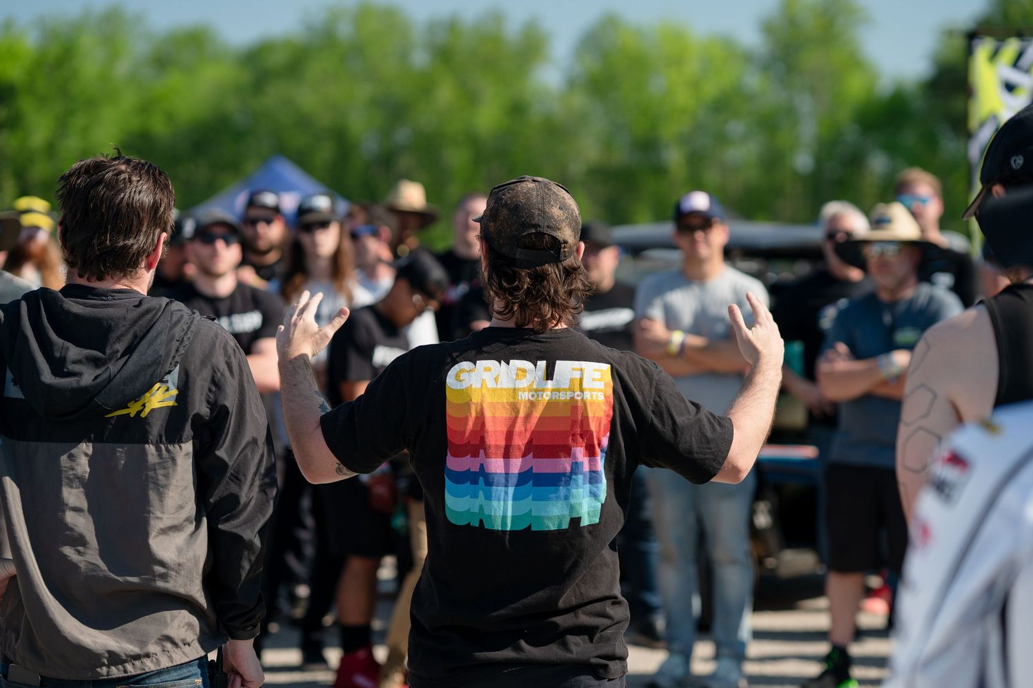 Group of motorsport enthusiasts gathered outdoors at a car event, one wearing a colorful GRDLife shirt.