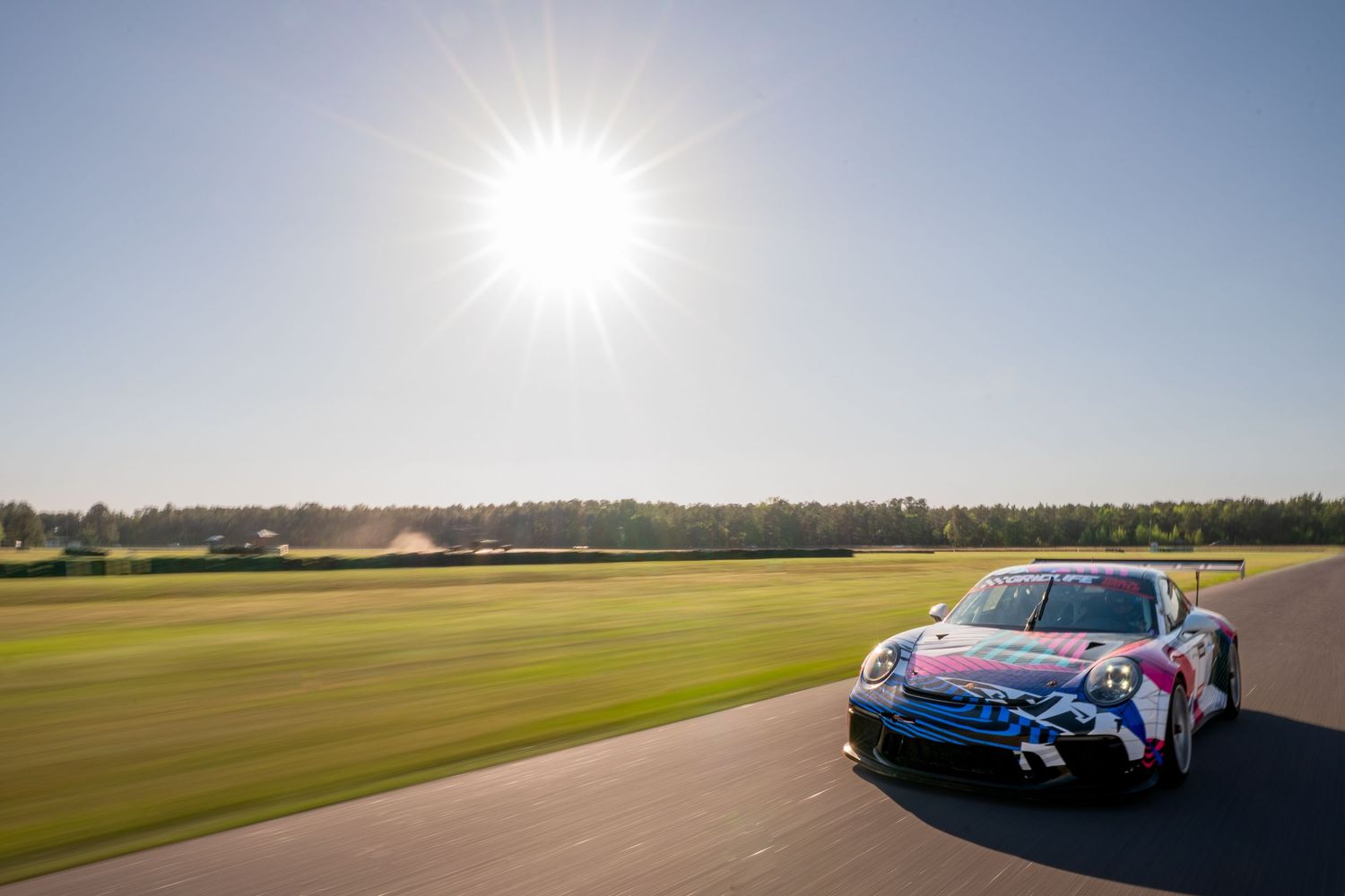 Colorful pink and blue race car speeding on a sunny track with bright sunburst and green treeline in background.
