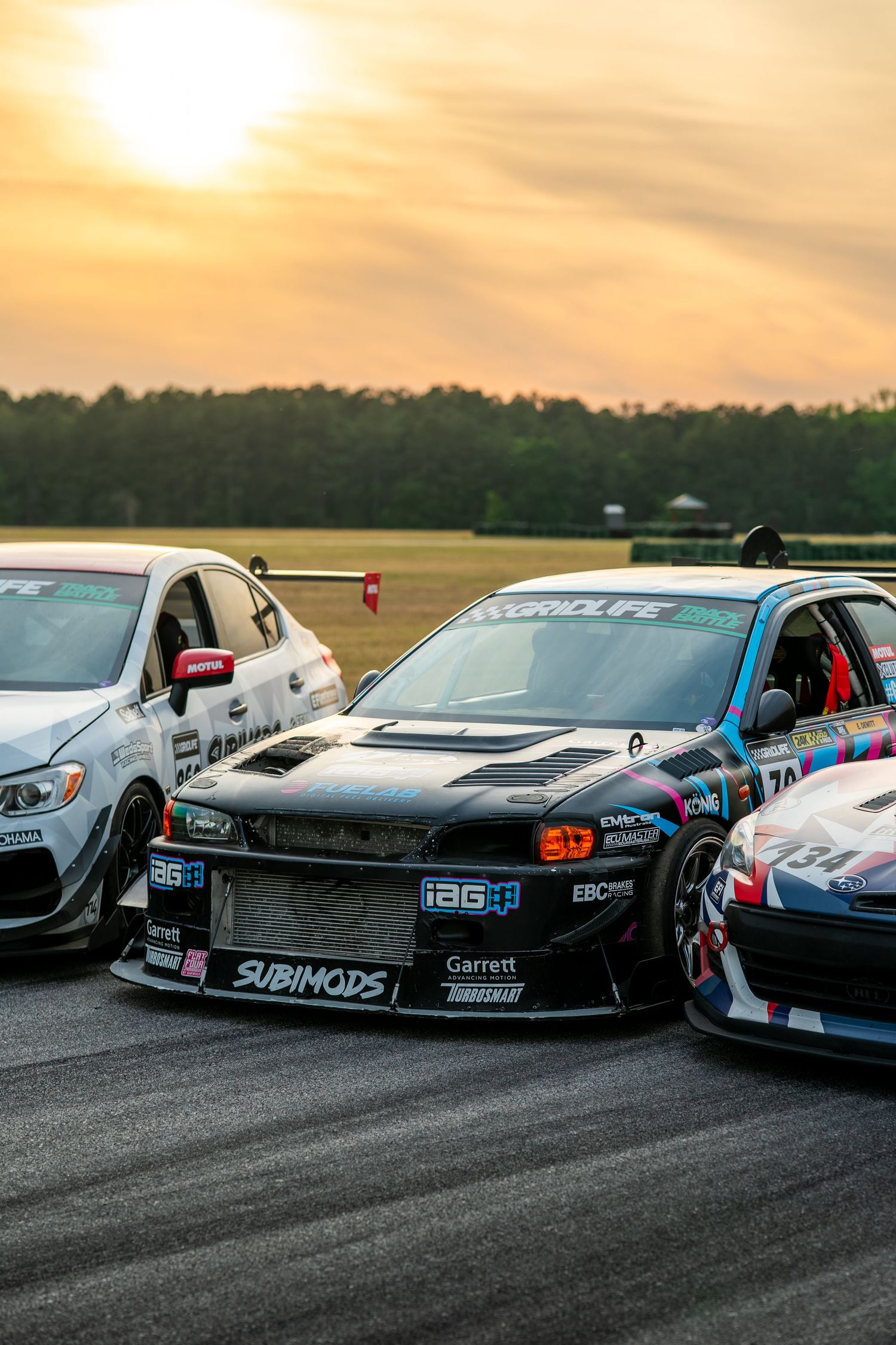 Three tuned race cars lined up on a track at sunset, including a black Subaru with IAG and Garrett sponsorship decals.