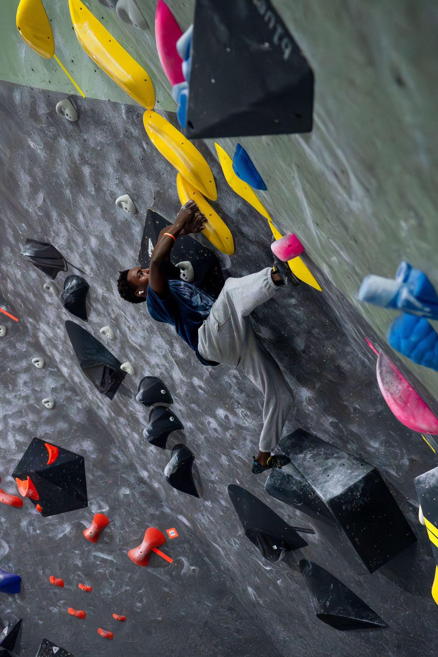 Climber in a blue shirt and gray pants scales a steep indoor bouldering wall covered with black, yellow, and orange holds.