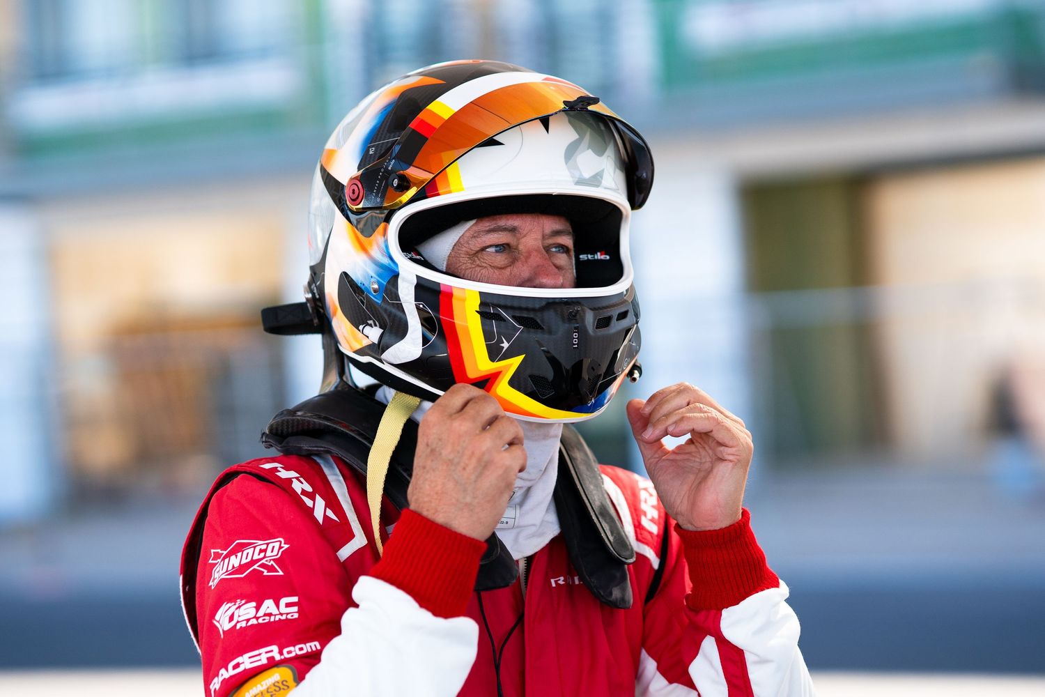 Racing driver adjusting orange and black helmet while wearing red racing suit.