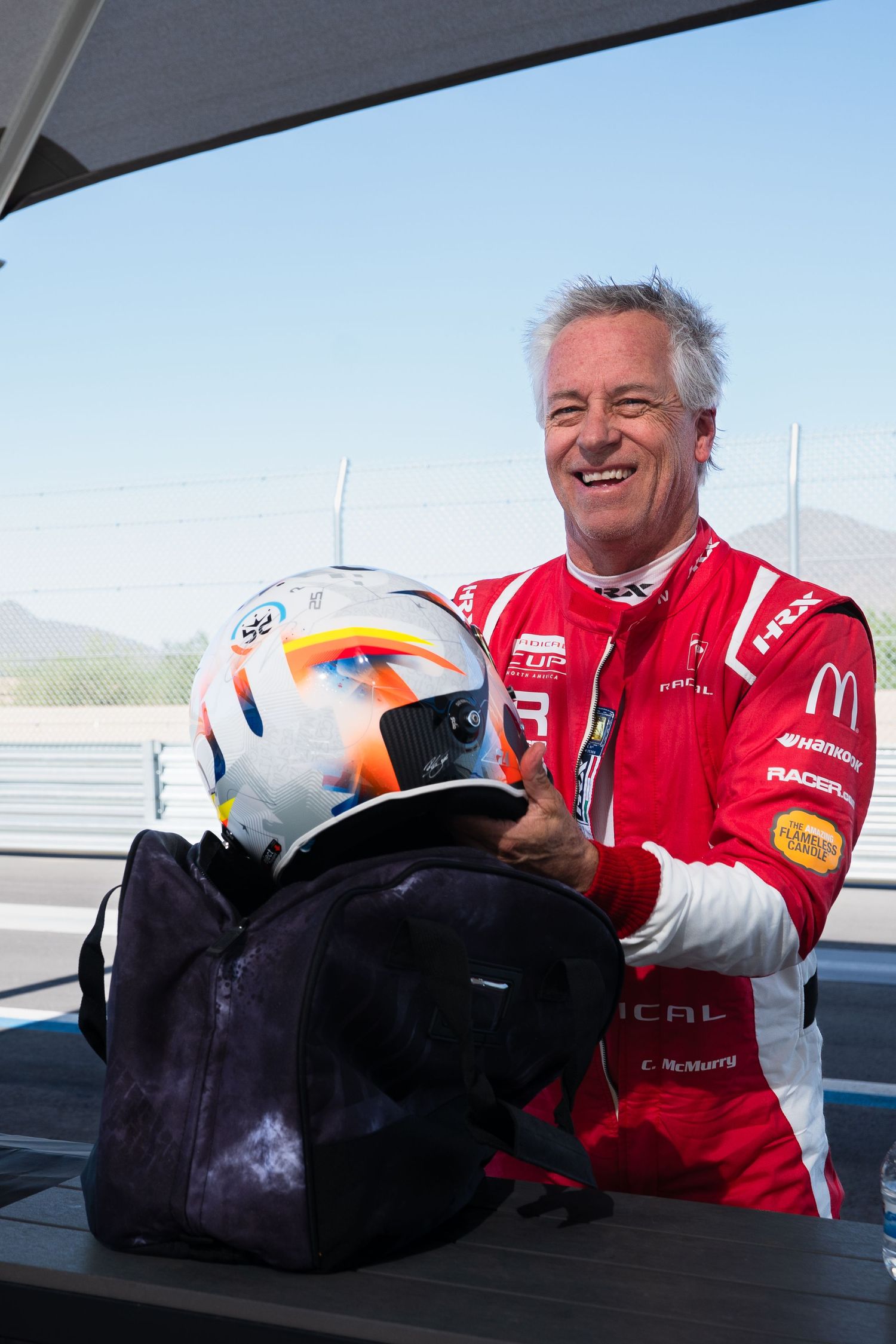 Person in red racing uniform examining and displaying protective racing helmets outdoors.