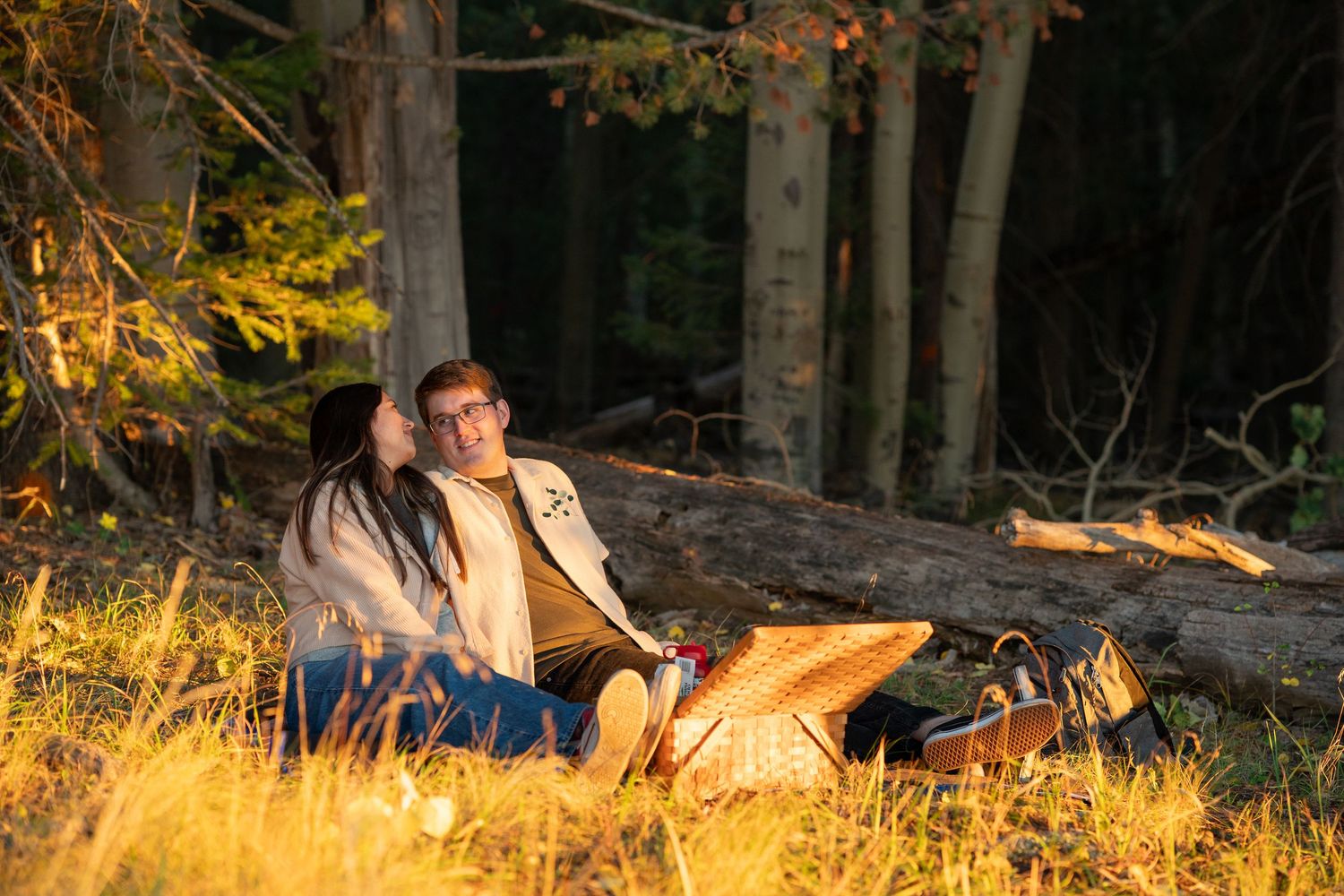 Couple sits close together on golden autumn grass beside a fallen log, enjoying a picnic with a wicker basket in a sunlit forest.
