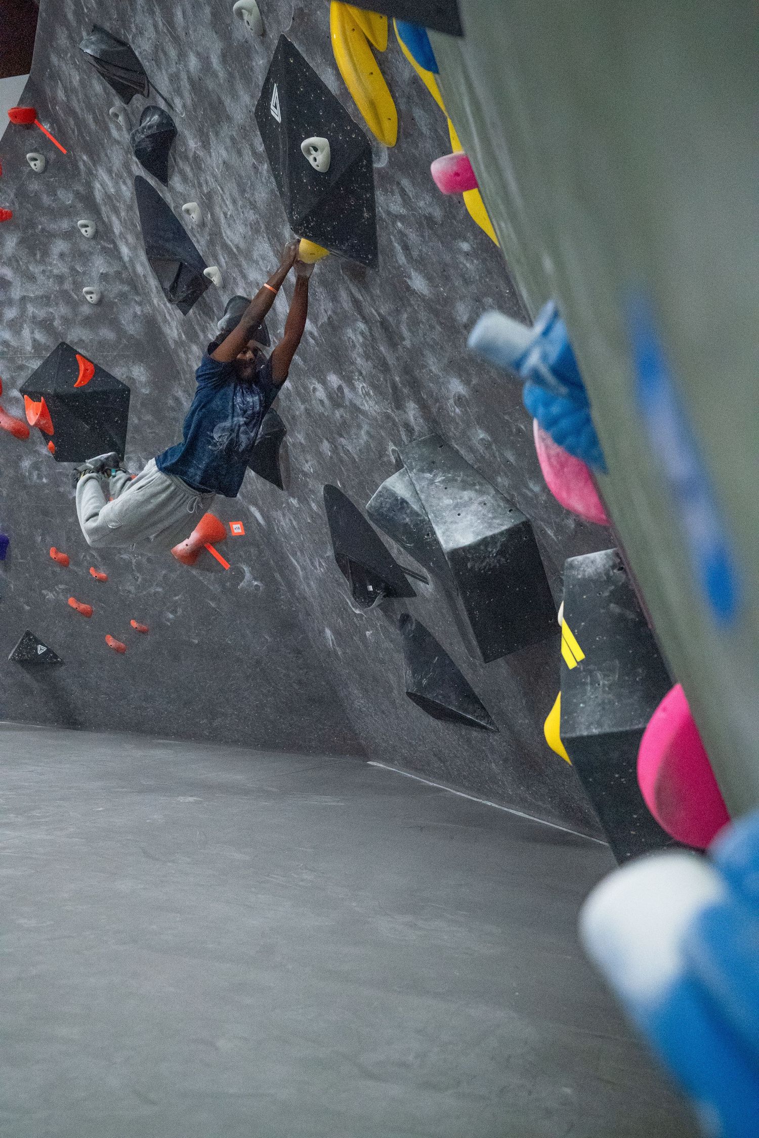 Climber hangs from a hold on an overhanging indoor bouldering wall covered in black, yellow and pink holds and volumes.