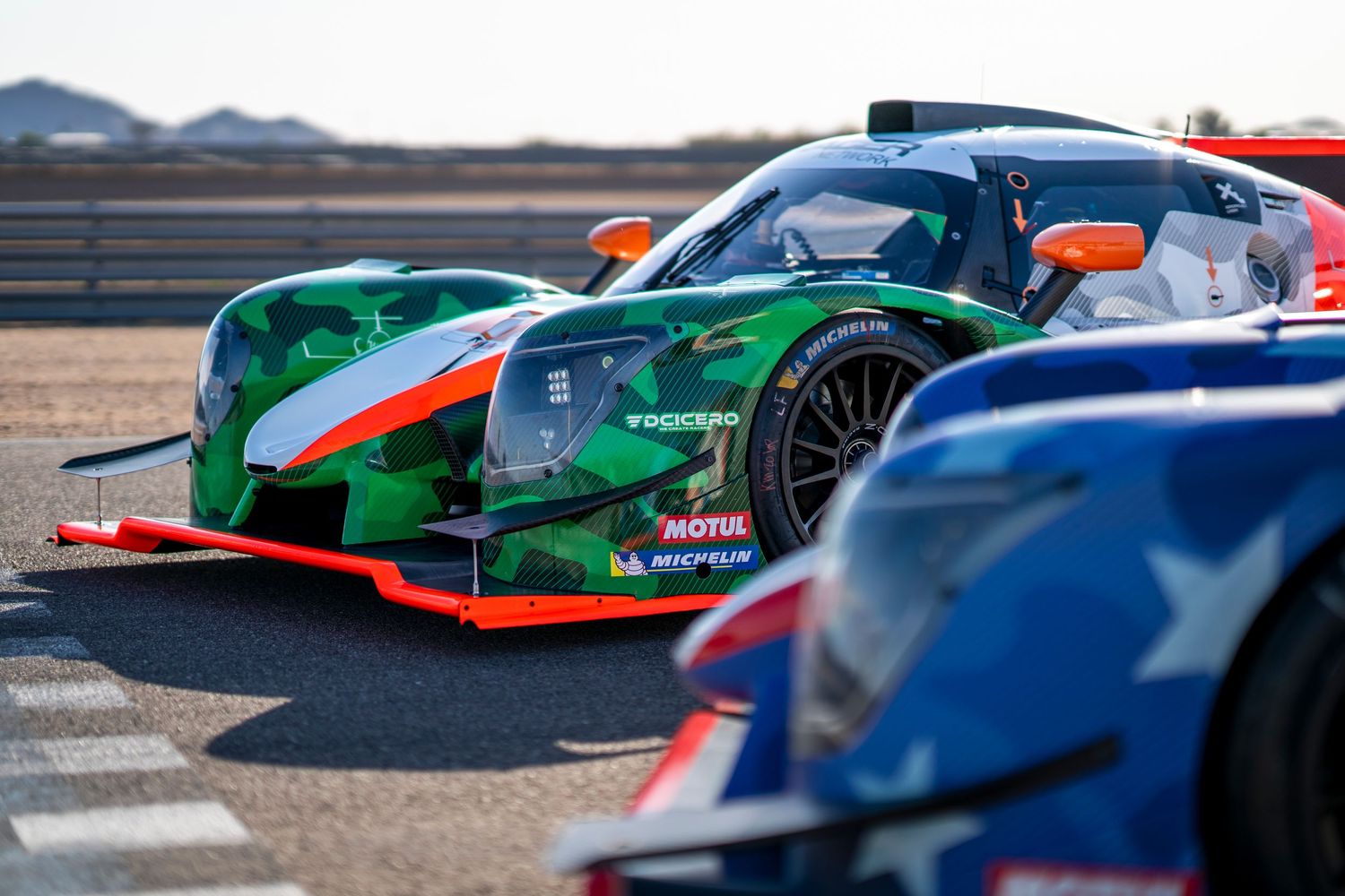 Sleek green and red racing prototype cars lined up on a racetrack under bright daylight.